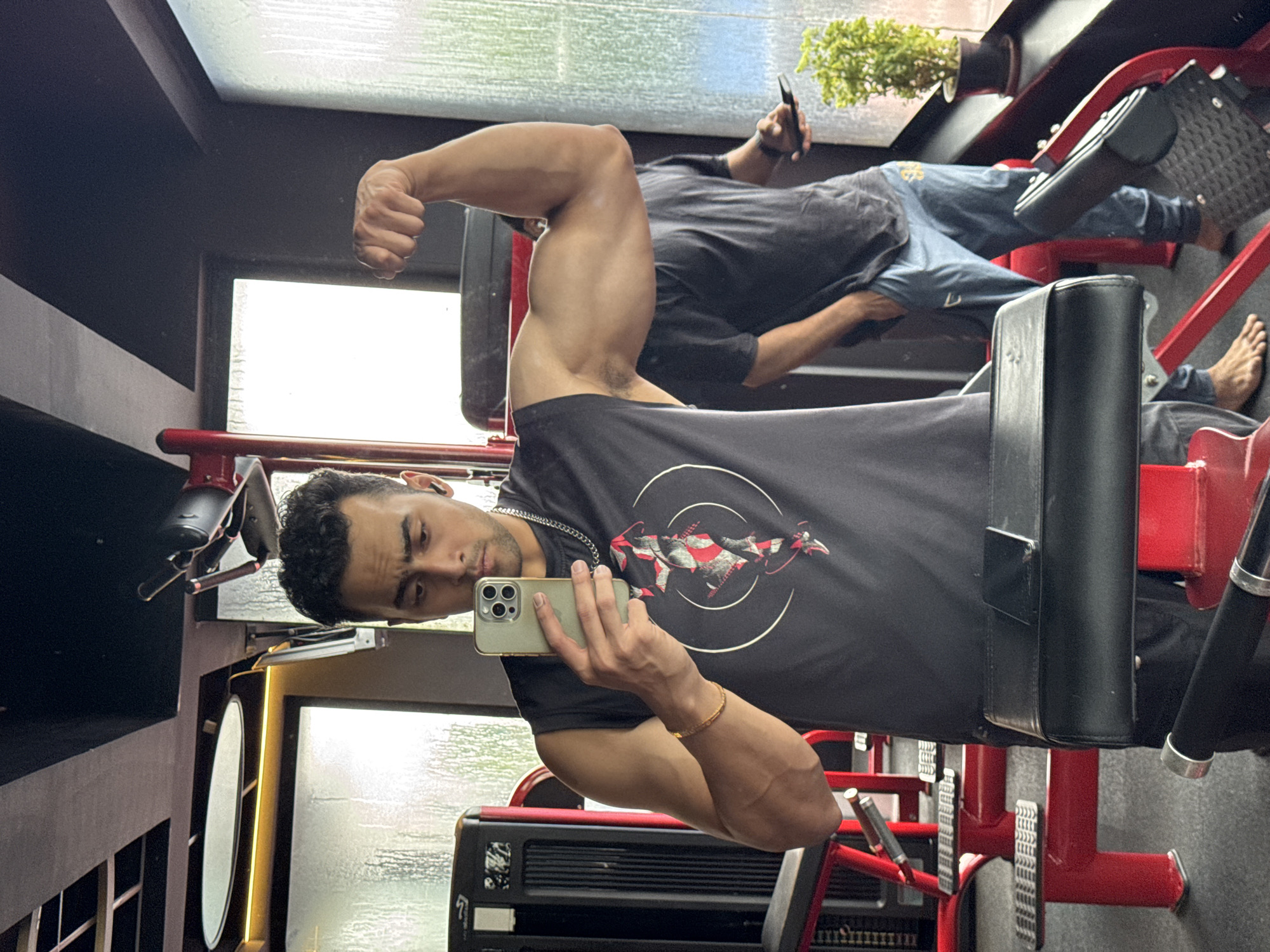 A man is taking a mirror selfie in a gym. He is wearing a black sleeveless shirt and black pants, and he is flexing his bicep. He is holding a light green smartphone in his left hand. In the background, another man is standing with his hands in his pockets, looking at his phone. The gym appears to be modern with red and black equipment and a large window with water droplets on it. The image captures a moment of fitness and self-expression in an urban gym environment. The presence of a specific gym in Kolkata, India, is not ascertainable from the provided image.