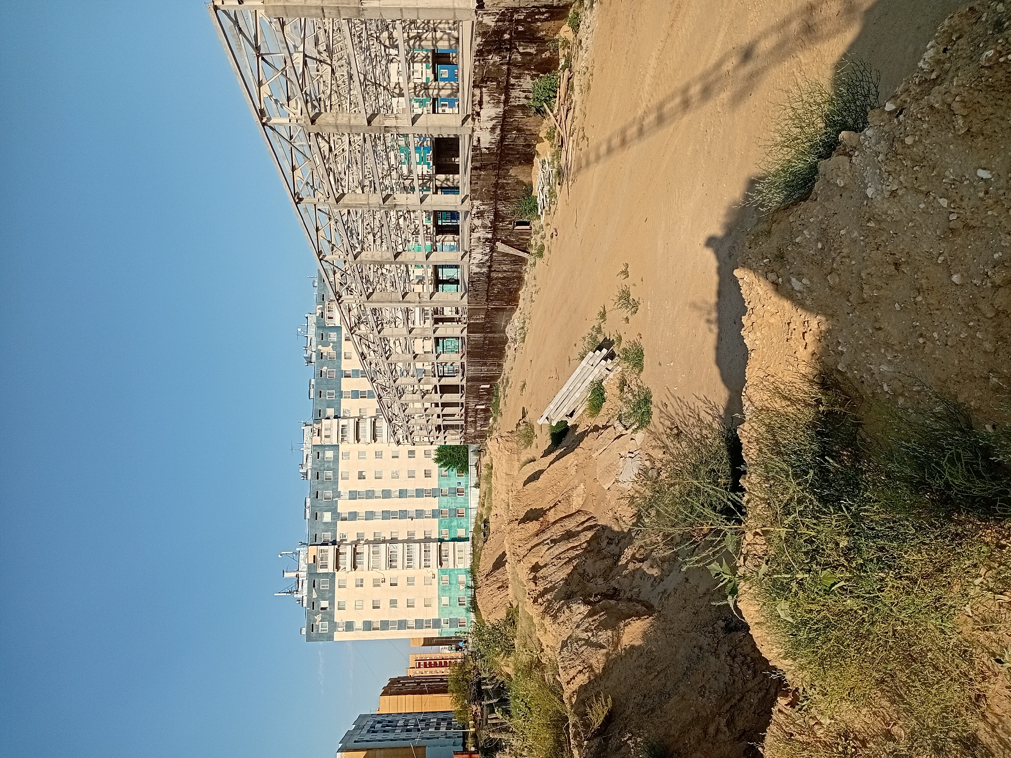 The image depicts an outdoor construction site in the city of Semey, Kazakhstan, under a clear blue sky. In the background, a large, multi-story apartment building with light-colored walls and a turquoise band across its upper floors stands prominently. Adjacent to the apartment building, to the right, is a partially constructed building with a visible metal and concrete frame. The foreground of the image is dominated by a dirt excavation, with uneven piles of sandy soil and sparse green vegetation growing on the mounds. Several stacks of lumber and some metal pipes are scattered around the site. The scene suggests an ongoing construction project in an urban environment.