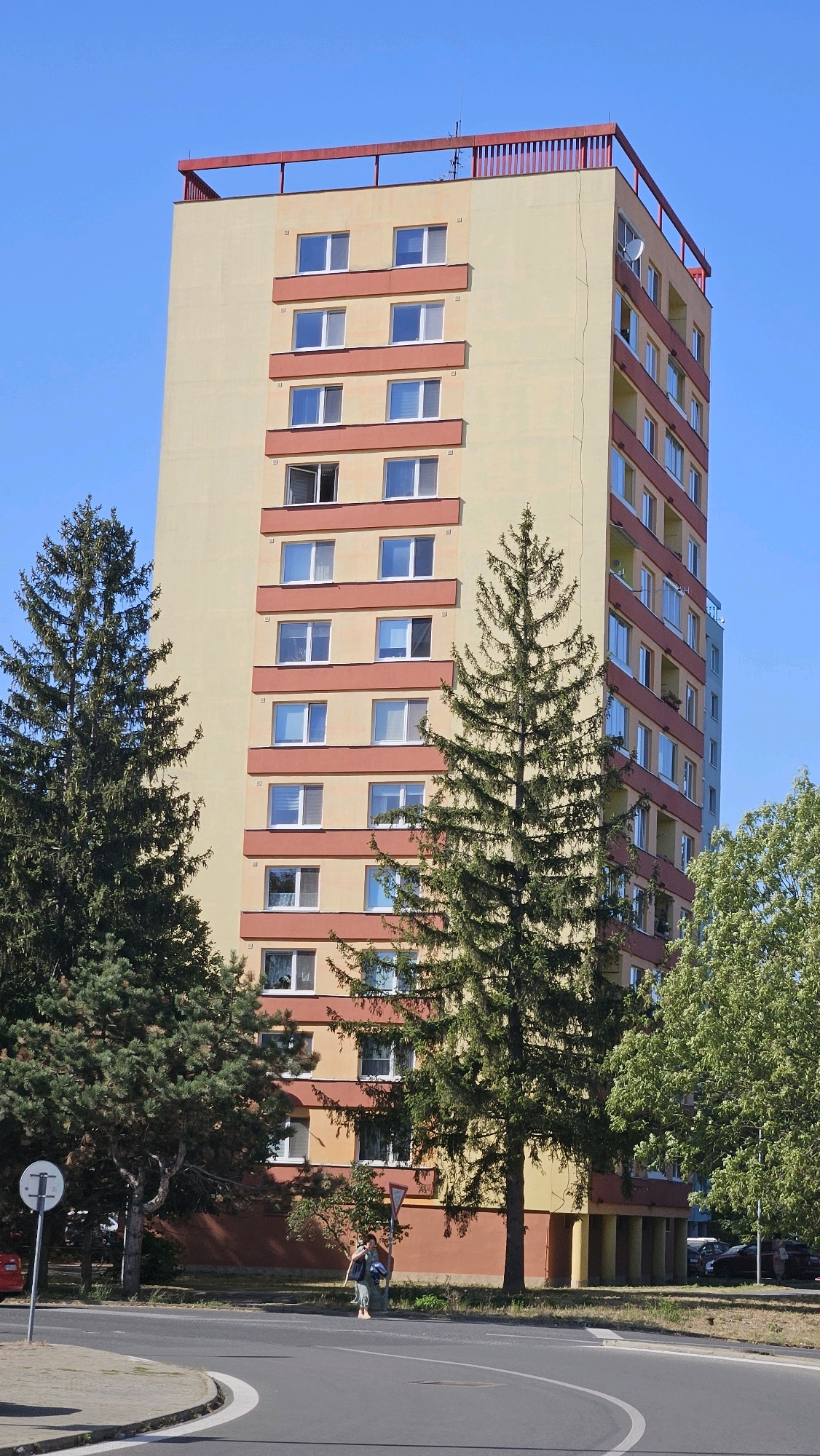 A tall, slender apartment building with a pale yellow facade and red trim is the primary subject of this outdoor, daytime photograph. The building has multiple stories, each with rows of windows. A red railing outlines the rooftop. Coniferous trees, one particularly tall and dense, stand in front of and to the side of the building. A paved road with white lane markings curves across the foreground, with a sidewalk and some greenery beyond. A solitary woman, dressed in light-colored clothing and carrying a bag, walks across the road. Several parked cars are visible behind the woman and to the right of the building, near a line of trees with green foliage. The sky is a clear, bright blue. The image appears to have been taken in a residential area of Prešov, Slovakia, given the architectural style and vegetation commonly found there.