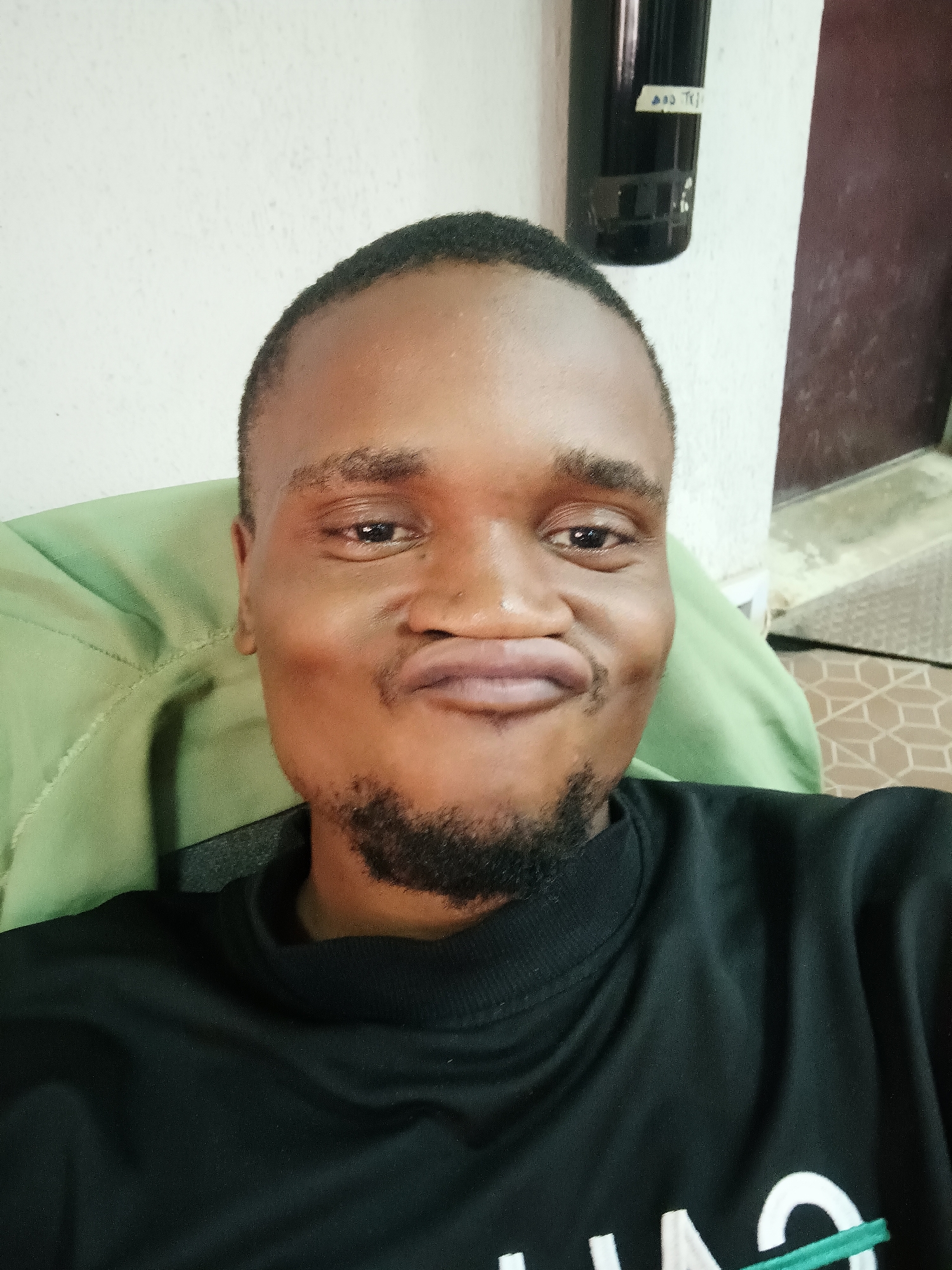 A close-up, front-facing selfie captures a young man with dark skin, short dark hair, and a neatly trimmed beard, seated in what appears to be a light green armchair. He is looking directly at the camera with a playful expression, puckering his lips into a pout. His eyes are bright and engaged. He wears a black long-sleeved shirt with partially visible white and green text or a logo on the lower right side, which seems to include letters like "AQ" and part of a "C" or "O" and a green diagonal line.
The setting is indoors, likely a casual living space in Kakuri Gwari, Nigeria. The background features a plain white wall on the left, with a dark, cylindrical object mounted on the wall in the upper right quadrant. This object has a light-colored label on it, showing legible text "TY" and what might be "100" or "200" before it, along with other smaller, less clear characters. To the far right, a reddish-brown doorframe or wooden panel is visible, and on the floor below, a brown and cream-colored patterned mat or rug can be seen. The lighting is even, suggesting an indoor setting with adequate illumination, possibly daytime or well-lit evening. The overall mood conveyed by the man's expression is lighthearted and engaging.