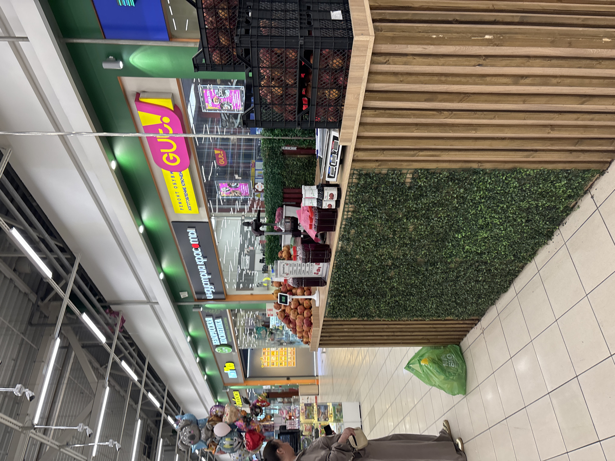 The image captures an indoor scene within a modern retail environment, likely a supermarket or shopping mall in Russia. The perspective is slightly elevated, looking down a wide aisle lined with various stores and product displays.
In the foreground, on the right, a person is partially visible, walking away from the camera. They are wearing a long, plain, light brown/grey robe or coat and light-colored shoes, and appear to be holding a small, light-colored bag or phone. A bright green plastic bag with "100% БИО" (100% BIO) written on it is lying on the white tiled floor near the person's feet.
To the right of the aisle, there's an elaborate display featuring a section of artificial green wall covering and wooden slatted paneling. Above this, a produce stand holds numerous fresh fruits, possibly apples or peaches, stacked in black crates and on a wooden table with a white weighing scale.
Along the left side of the aisle, multiple storefronts are visible with prominent signs and bright lighting. One large sign features the bold pink and yellow text "GU!", with smaller Russian text below it reading "РЕМОНТ ОБУВИ ИЗГОТОВЛЕНИЕ КЛЮЧЕЙ" (SHOE REPAIR KEY MANUFACTURING). Other signs, predominantly with a green background, include "ТВОЯ АПТЕКА" (Your Pharmacy) and "БАЛАУНДРИЯ КРАСОТЫ" (Beauty Laundry - likely a store name). Further down the aisle, various other retail displays and a collection of metallic and colorful balloons are visible.
The setting is brightly illuminated by numerous overhead fluorescent strip lights, characteristic of a commercial space. The overall impression is one of a clean, organized, and busy shopping area.