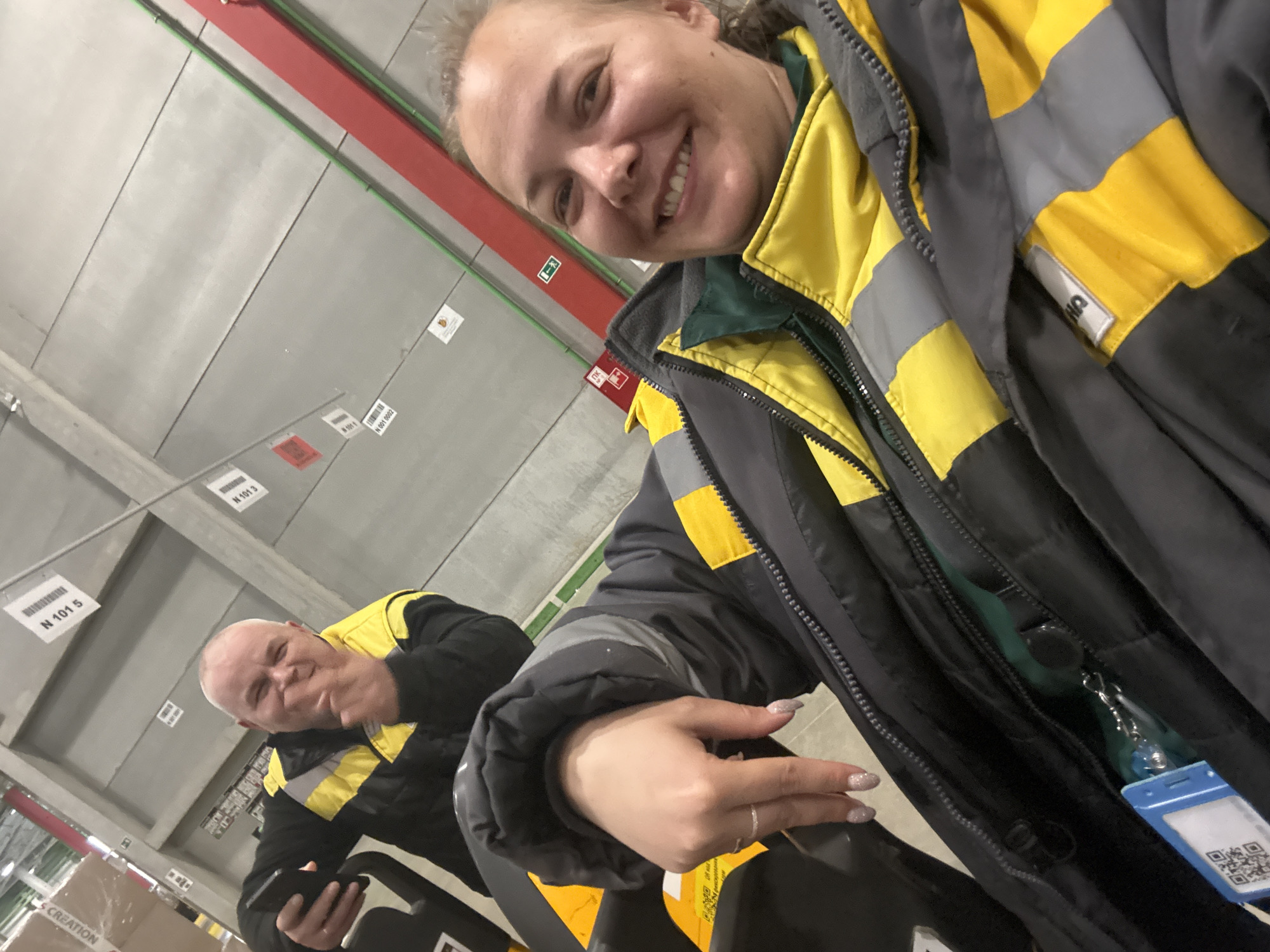 Two individuals are visible in this selfie, possibly taken inside a large warehouse or industrial facility. The individual in the foreground, likely the one taking the photo, is a fair-skinned woman with a bright smile. She's wearing a dark-colored jacket with yellow and gray reflective stripes. Her fingernails appear to be decorated with glitter or a light-colored polish. Hanging from her jacket zipper is a blue badge or identification card. In the background, to the left, a second person, an older man with short gray hair, is visible. He is also smiling and wearing a similar black and yellow striped jacket.
The environment consists of high ceilings with a gray industrial finish. Various overhead signs or labels, such as one labeled "N 101 5," are suspended from the ceiling. Other ceiling fixtures and piping are visible, including green and red piping. The warehouse facility contains shelving or storage racks in the distance, with some boxes and materials visible on the lower levels.
There is no indication that the photo was taken in Lupanovo, Russia. The location cannot be confirmed from the image.