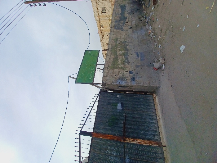 This image depicts an outdoor scene in the city of Kafr Sousa Municipality, Syria, focused on a gateway and an adjacent wall under an overcast sky.
In the foreground, the ground transitions from a lighter concrete/asphalt surface on the left to a dirt and debris-strewn area on the right, littered with various pieces of trash including paper and small rocks.
On the left, a large, dark-colored, industrial-style metal gate, approximately two meters tall, is partially open. The gate features a grid pattern and an ornate, spiked top section. Rust is visible on the gate's frame and hinges. The gate is connected to a sturdy concrete pillar on its right side.
To the right of the gate, extending further right across the midground, is a low, unpainted concrete block wall, roughly constructed and showing significant weathering, discoloration, and grime. Patches of darker stains and possibly moss are present on its surface.
Mounted above the concrete wall and pillar, a rectangular green sign with white or light-colored Arabic text (mostly illegible due to resolution and angle) is visible. The sign is supported by metal poles and is angled upwards slightly.
In the background on the right, a two-story building constructed of light-colored masonry (possibly stone or concrete blocks) is visible. It features multiple windows and appears to be in a state of disrepair or aged. Further left in the background, behind the open gate, another structure made of similar materials is partially visible.
Overhead, a network of utility wires spans the scene. A dark brown wooden utility pole with several crossarms and multiple power lines runs vertically along the far right edge of the frame. A prominent dark wire sags horizontally across the upper-middle section of the image. The sky is a uniform, bright grey, indicating an overcast day. No individuals are present in the scene.