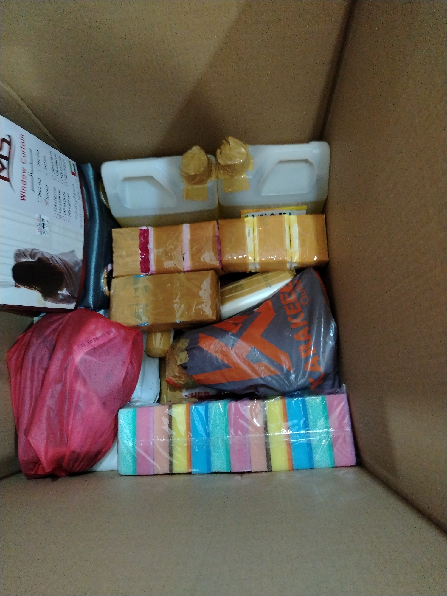 The image shows the inside of a cardboard box containing various items. The box appears to be in a warehouse or storage area. Among the items are several packages wrapped in brown paper and tape, some plastic containers with yellow lids, a red plastic bag, and a package of colorful sponges. There is also a piece of paper or cardboard with text and an illustration of a person. The scene is well-lit, and the objects are arranged in a somewhat cluttered manner within the box. There is no discernible information within the image to indicate a specific location in Dubai, United Arab Emirates.