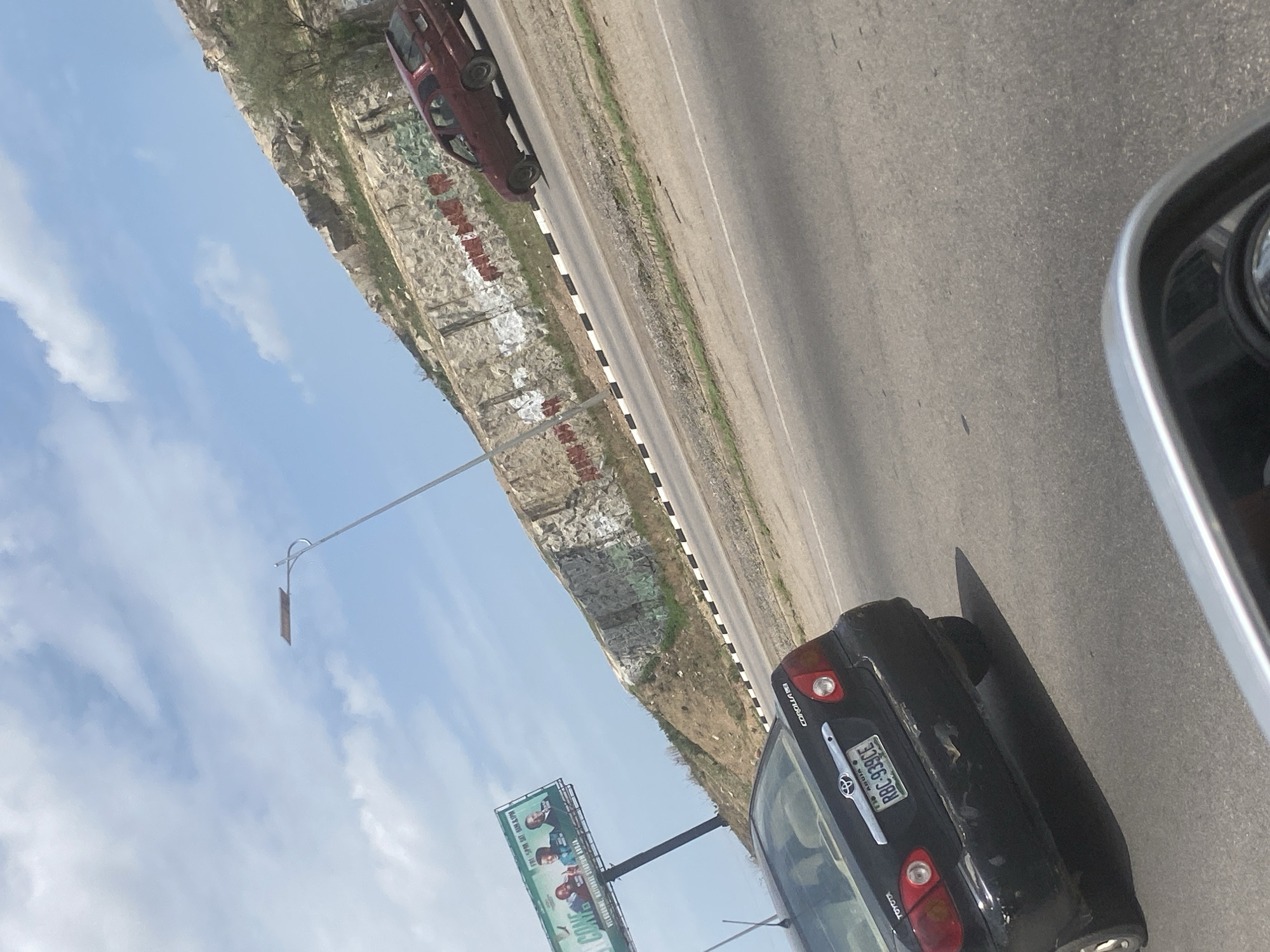 The image depicts a roadside scene in what appears to be the city of Abuja, Nigeria. The view is from inside a vehicle, with part of the side mirror visible. A black Toyota Corolla is in the adjacent lane. The number plate reads "RBC-939CE" and identifies the location as Abuja.
A large billboard is seen to the left, displaying images of individuals, presumably related to a political campaign, along with the text "LET'S COME TOGETHER, FOR BETTER TOMORROW".
The road is bordered by a rocky embankment painted with graffiti. Another vehicle, possibly red, is seen parked on the embankment. A street light is visible extending over the road. The sky is partly cloudy.