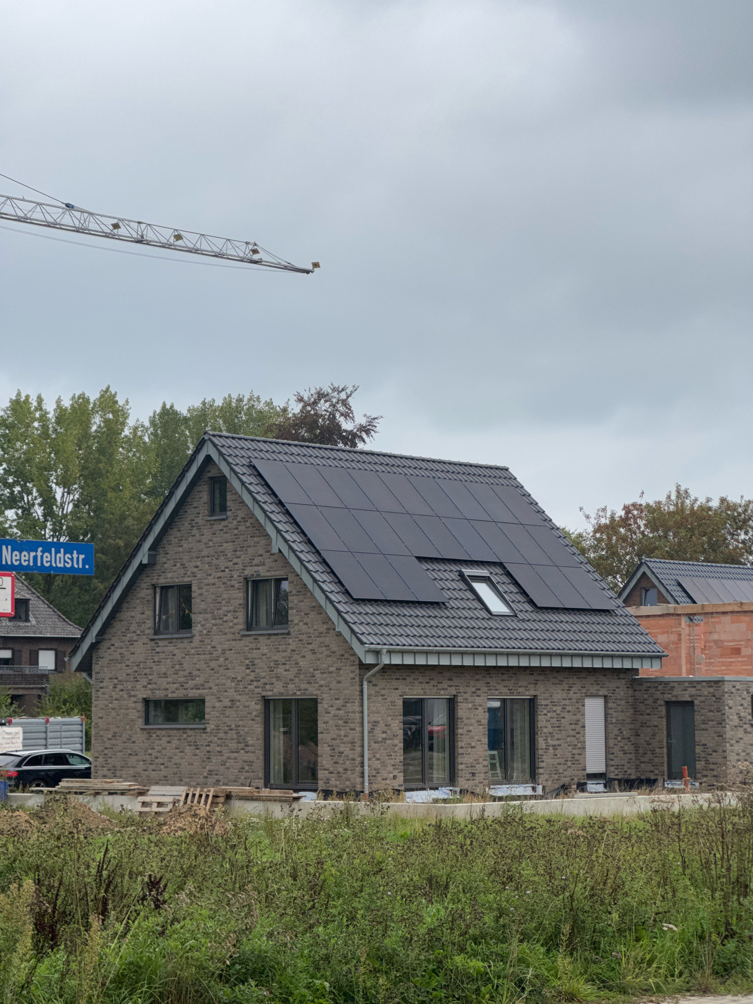 The image shows a street scene in Cleves, Germany, featuring a modern brick house with solar panels on its roof. A street sign reads "Neerfeldstr." A construction crane is visible in the sky, suggesting ongoing development in the area. The house has large windows and a dark-tiled roof. In the foreground, there is dense green vegetation. A dark-colored car is parked to the left of the house, near a construction trailer. To the right of the main house, another building is under construction, with visible brickwork and scaffolding. The sky is overcast.