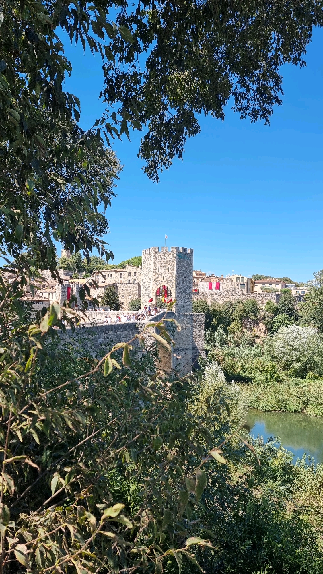 The image shows a view of the medieval bridge and castle of Besalú, Spain. The stone bridge, with its characteristic arches, spans a tranquil river. A prominent stone tower, part of the castle complex, stands on the bridge. Red banners adorn the castle walls, suggesting a festive occasion or historical reenactment. People are visible on the bridge, some appearing to be participating in the event. The surrounding landscape is lush and green, with trees and foliage framing the scene and a body of water reflecting the clear blue sky. The architectural style and the river suggest the specific location is Besalú, a well-preserved medieval town in Catalonia, Spain.