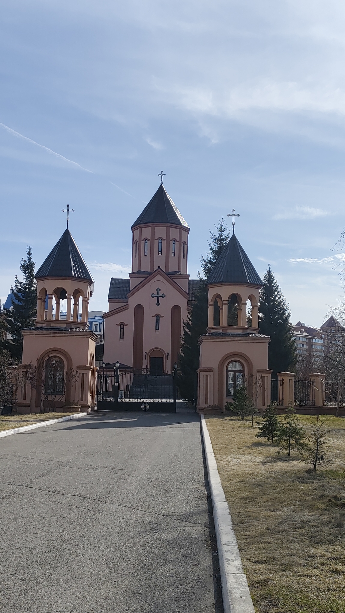 This image captures an Armenian Apostolic church in Krasnoyarsk, Russia, under a bright, clear sky with scattered high clouds. The church, constructed with a light reddish-brown or peach-colored facade and dark roofs, features prominent architectural elements characteristic of Armenian design.
The main building has a central, multi-sided drum topped by a steep, dark conical roof, which is crowned with a cross. Below the drum, the main structure displays a large cross relief above its entrance, and narrow, tall windows. Two smaller, bell tower-like structures flank the main church building, positioned at the end of a paved driveway. These pavilions also have dark conical roofs topped with crosses and feature arched openings, suggesting they might house bells or serve as entry points.
A black, ornate metal gate stands closed, blocking the path to the church entrance. A wide asphalt driveway leads directly to this gate, with concrete curbs delineating the edges of the pavement. On both sides of the driveway, there are patches of dry grass and several small evergreen trees or shrubs. Taller pine trees are visible around and behind the church structures, and other buildings can be discerned in the background beyond the church grounds.
The scene depicts a peaceful setting, likely during daylight hours given the bright illumination. No people or explicit text are visible in the photograph. The overall impression is one of a well-maintained religious complex.