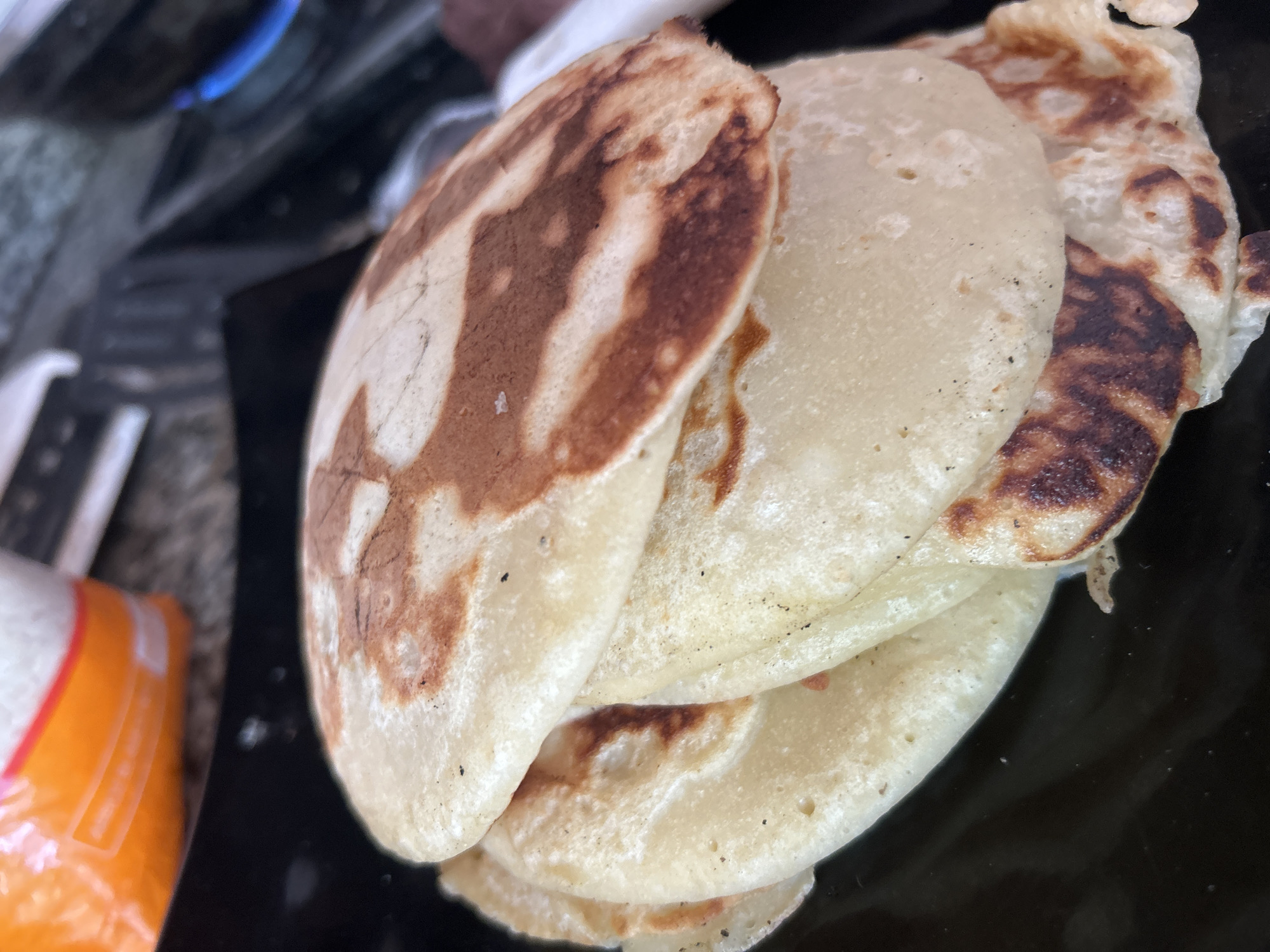 A close-up, overhead shot shows a stack of golden-brown and pale pancakes on a black plate. The pancakes appear to be slightly unevenly cooked, with some areas darker than others, indicating they were made on a griddle. The texture of the pancakes is visible, with small air bubbles on their surface. In the background, out of focus, is a bright orange and white package, possibly containing more ingredients or food. The scene is lit with natural light, casting subtle shadows. There are no people or animals visible in the image. The location is identified as Santa Ana, Venezuela.