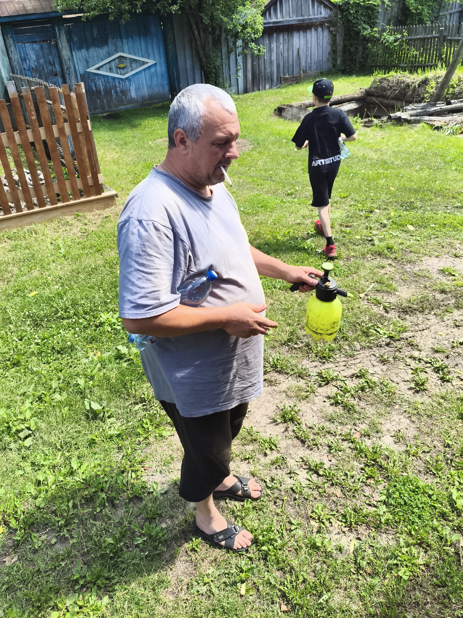 In a rural backyard, likely in the village of Ilmino, Russia, a middle-aged man with graying hair stands in the foreground, smoking a cigarette and holding a yellow spray bottle in his right hand. He is wearing a light blue t-shirt and dark pants. To his left, a partially filled plastic water bottle is tucked into his waistband. In the background, a young boy wearing a black t-shirt with "ARTSTUDIO" printed on the back, black shorts, and a baseball cap runs away from the camera. He is carrying another plastic water bottle. The yard is covered in green grass and patches of dry, cracked earth. A rustic wooden fence and a weathered blue gate are visible in the background.
