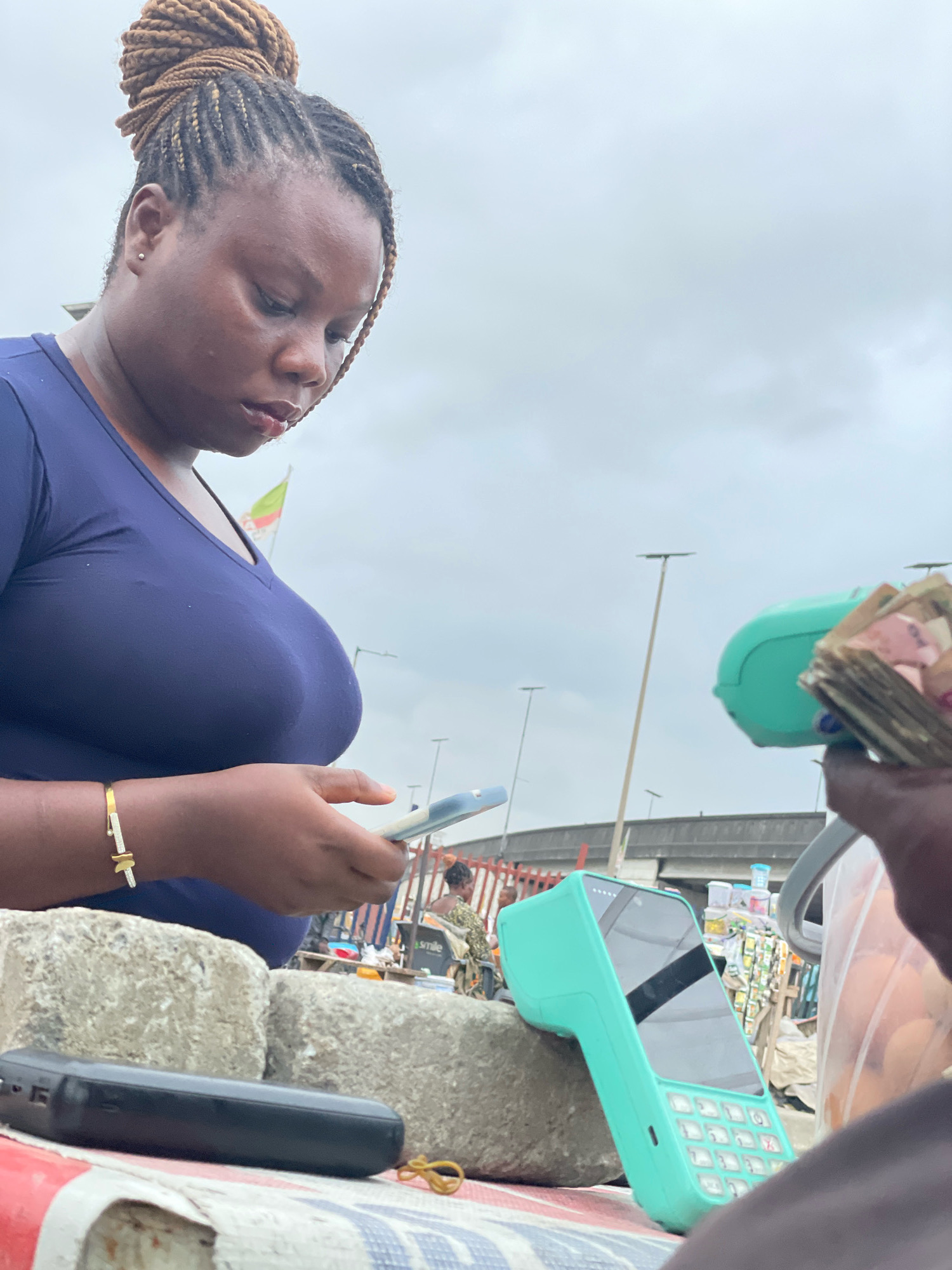 A close-up, eye-level shot captures a woman in Ajah, Nigeria, with a dark complexion and her hair styled in intricate braids pulled into a bun. She wears a dark blue V-neck shirt and a gold and diamond bracelet on her left wrist. The woman is looking down, seemingly engaged with a smartphone in her hands, which are positioned over two large, rough-hewn grey stones. Beside the stones, on a surface covered with a striped cloth, lies a black electronic device and a yellow rubber band.
In the background, to the right of the woman, a teal-colored point-of-sale (POS) device rests on a stone. A hand is visible holding a fan of cash in front of the POS device. Further back, out-of-focus figures and stalls suggest a market environment, with goods displayed on shelves and a glimpse of a green and white flag. Streetlights and an elevated structure are visible against a cloudy sky, indicating an outdoor urban setting. The overall impression is that of a transaction taking place in a busy marketplace.
