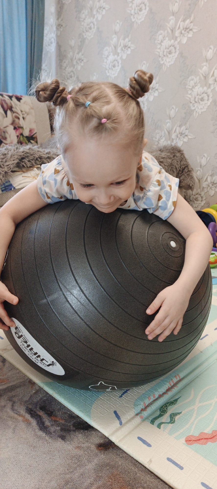 A young child, likely a girl, is pictured lying belly-down on a large, dark gray exercise ball in an indoor setting in Chaikovsky, Russia. The child has light-colored hair styled into two small buns on top of her head, secured with pink and light blue elastic bands. She is wearing a short-sleeved top patterned with small blue and gold heart shapes. Her head is tilted downwards, and she appears to be smiling and focused on the ball, gripping it with both hands.
The exercise ball, which features concentric grooves, has white text visible on its surface, including "LATEX BAND," "TINKER BODY," and "DIA 55CM." A partial brand name, possibly "SHIRIE" or similar, is also present.
The background consists of light-colored wallpaper adorned with a subtle white and gray floral pattern. To the left, a portion of a patterned sofa or cushion is visible. The child and the ball are situated on a light blue and white playmat featuring various colored abstract shapes and lines, which lies over a patterned carpet. A glimpse of colorful toys can be seen in the background to the right. The scene is brightly lit, suggesting it is daytime. The child's expression conveys joy and engagement in the activity.