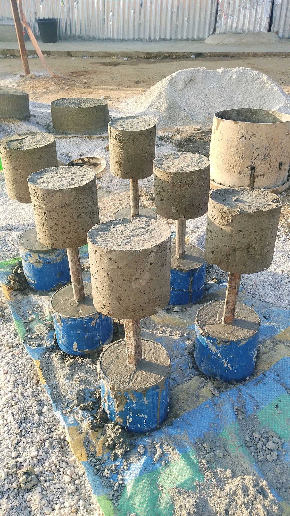 The image depicts a construction site in Indo, Nigeria. The scene is characterized by a collection of cylindrical concrete samples, each mounted on a metal rod and resting on a blue tarp. These samples appear to be test specimens for construction materials. Surrounding the samples are piles of gravel and sand, typical of a building or infrastructure project. In the background, a corrugated metal fence is visible, suggesting a secured perimeter. The overall environment indicates an active outdoor construction area.