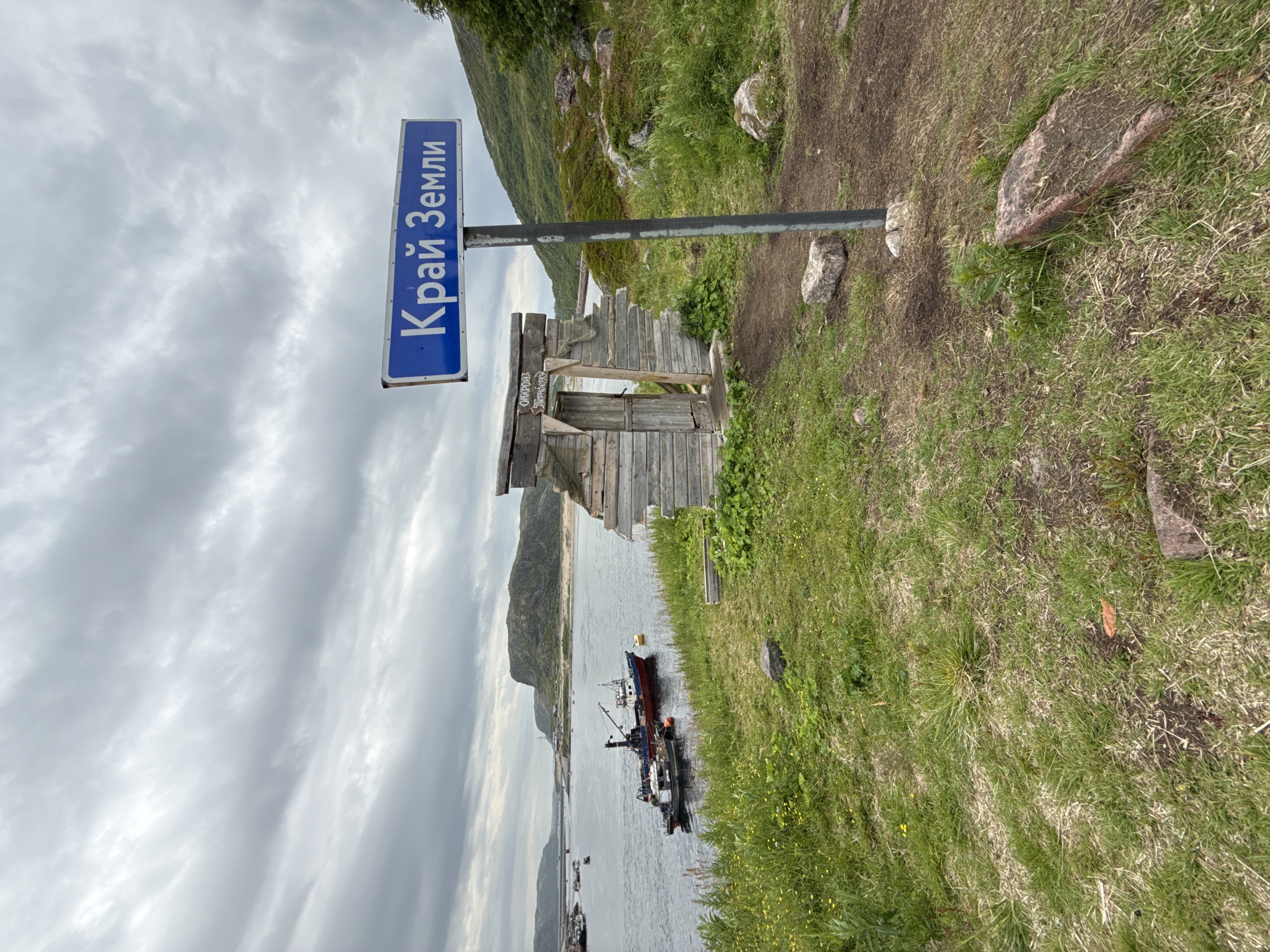 In Teriberka, Russia, a weathered wooden structure resembling a small, abandoned shack stands on a grassy hill overlooking a bay. A blue sign with white Cyrillic lettering, reading "Край Земли" (Krai Zemli), meaning "Edge of the Earth," is mounted on a metal pole in front of the shack. The shack has a simple wooden door and appears to be in disrepair, with gaps in its wooden walls. In the background, several fishing boats are docked or anchored in the water. The landscape is rugged, with rocky hills and sparse vegetation under a cloudy sky. The scene evokes a sense of remoteness and the end of the world.
