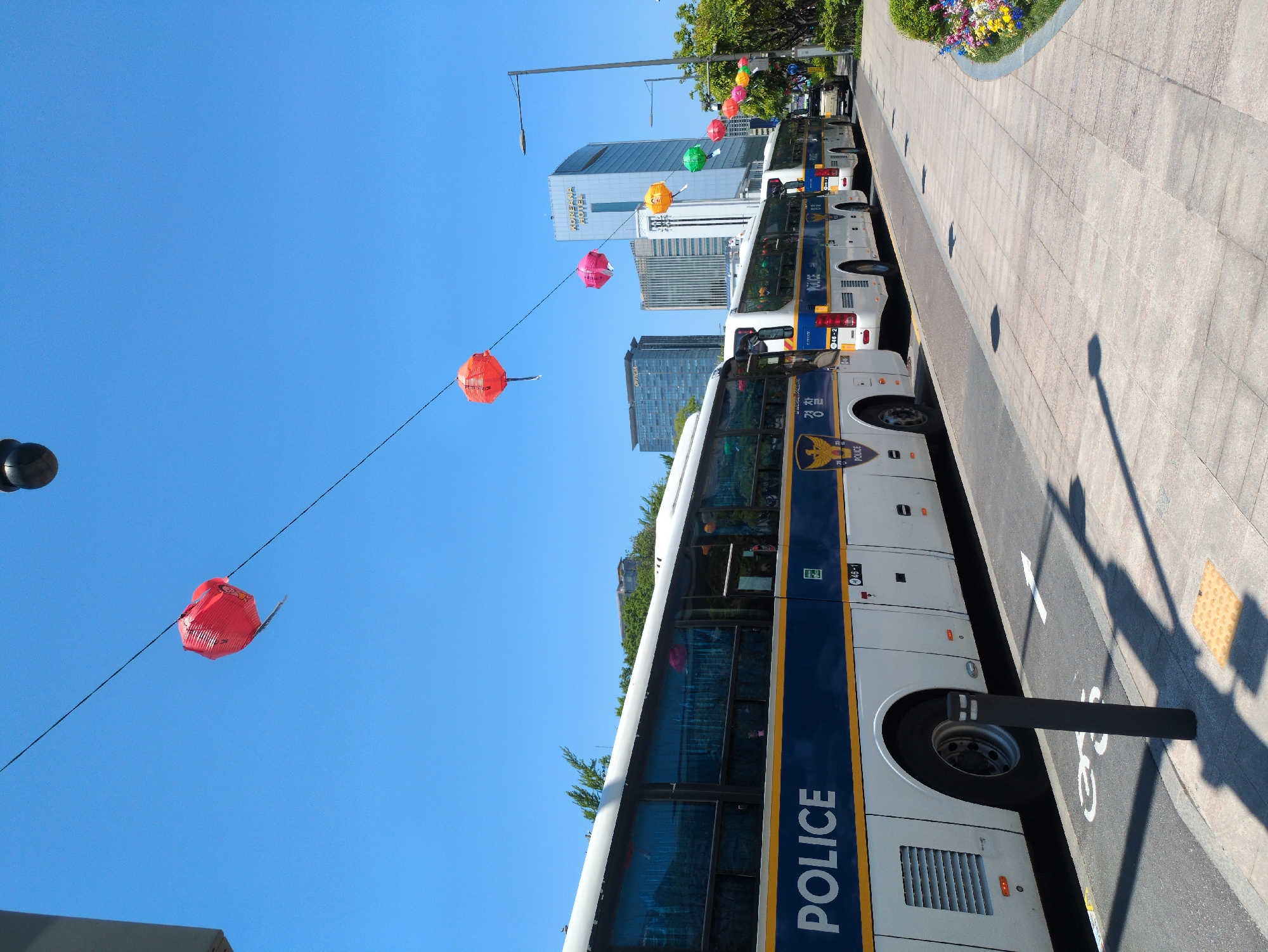 The image depicts a street scene in Seoul, South Korea, characterized by a row of police buses parked along a pedestrian walkway. The buses are white with a dark blue stripe and prominently display the word "POLICE" in yellow lettering. Lanterns, varying in color from red and orange to pink and green, are strung overhead. In the background, buildings including the Koreana Hotel, are visible against a clear blue sky. The walkway is paved and features flowerbeds at its edge. A street lamp and bollard cast shadows. A bike lane marking is visible on the street. The image is taken at an oblique angle.