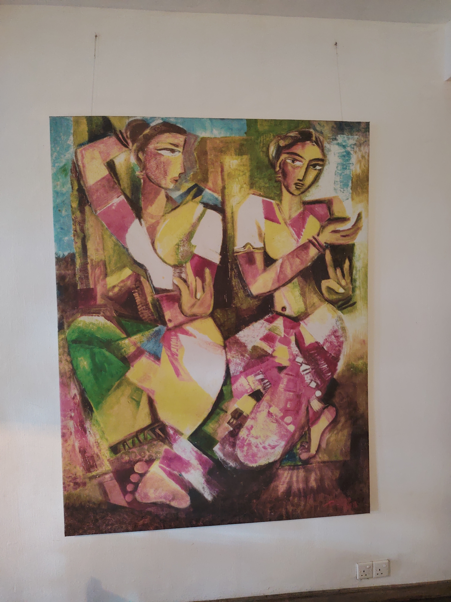 A framed painting hangs on a white wall, suspended by a thin wire. The painting depicts two figures, likely women, rendered in a bold, cubist-inspired style with broad brushstrokes and a vibrant color palette dominated by yellows, pinks, greens, and blues. The figures are depicted in a seated or kneeling pose, with their bodies fragmented into geometric shapes. Both figures are wearing traditional Indian attire, including ornate skirts and blouses. The figure on the left has her arm raised and bent behind her head, while the figure on the right has her hands positioned in a gesture of offering or blessing. The background of the painting is abstract and textured, with swirling colors and forms. Below the painting, two white electrical outlets are visible on the wall, and a dark wooden baseboard runs along the bottom of the wall. The lighting suggests an indoor setting with ambient light.