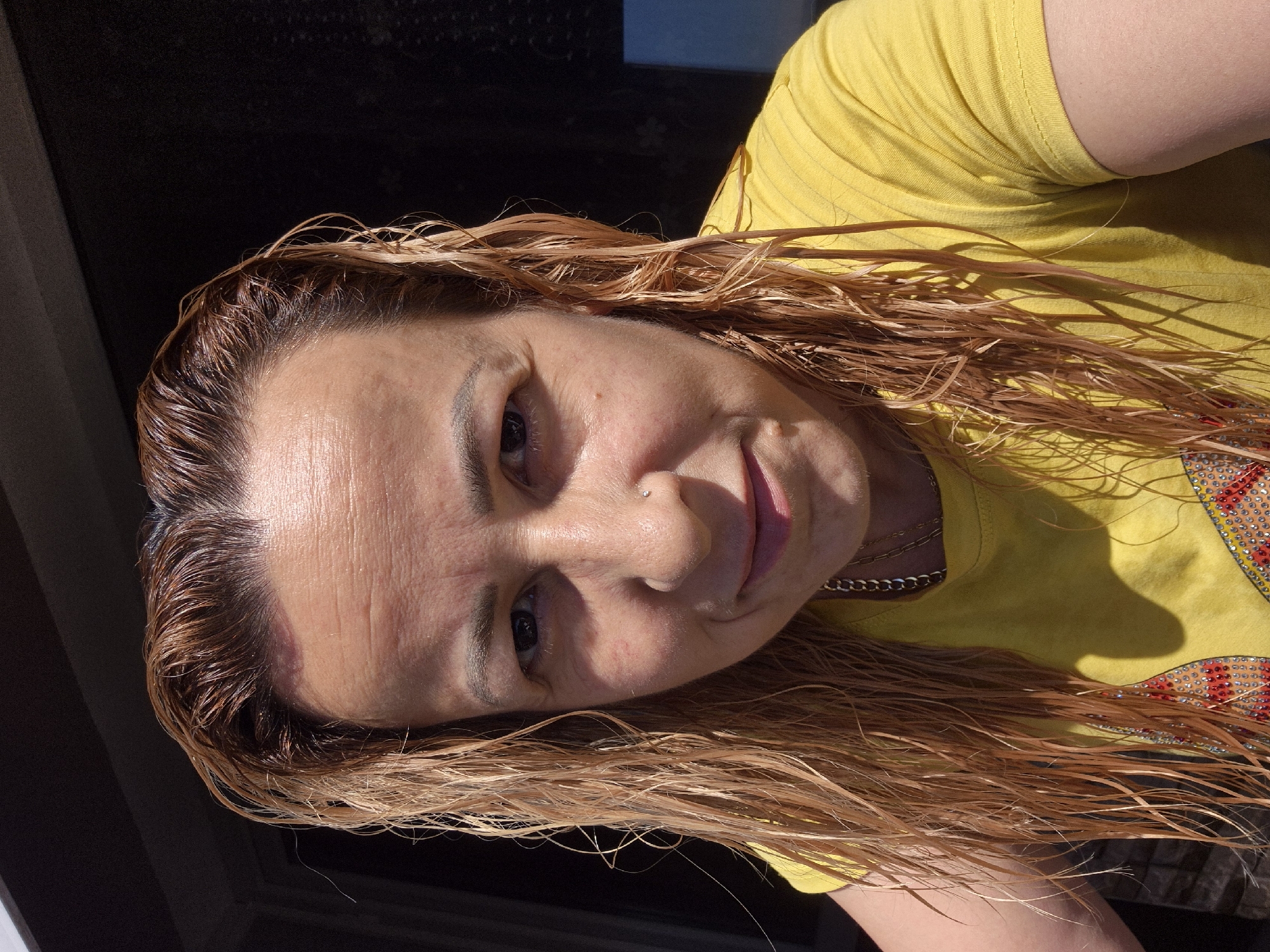 A woman with blonde, wet hair is shown from the chest up, looking at the camera. She is wearing a yellow t-shirt with a colorful design on the front. Her hair is parted in the middle and falls past her shoulders. She has a nose ring and appears to be outdoors, with sunlight casting shadows on her face and shirt. The background is dark and out of focus, with a hint of a window or door frame visible on the left side of the image.