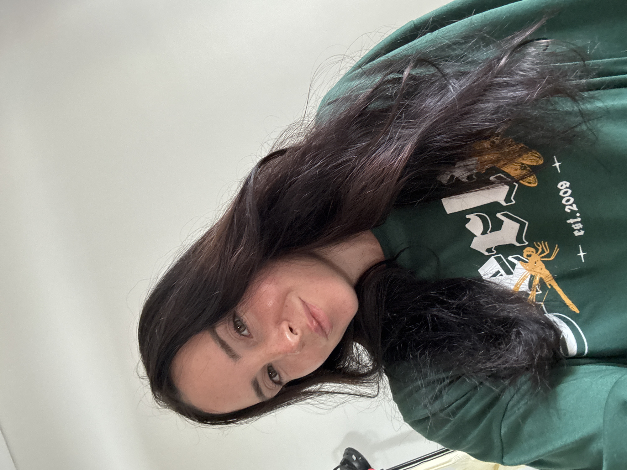 A medium-shot, eye-level, indoor photograph shows a woman from the chest up, looking directly at the camera. She has long, dark brown hair that falls over her right shoulder and is partially obscuring her face. Her hair is slightly windswept. The woman has fair skin with freckles, brown eyes, and is smiling slightly. She is wearing a dark green sweatshirt with white and orange graphics, including a dragonfly and text that reads "est. 2009". The background is a plain, light-colored wall. The lighting is bright and appears to be natural light from a window. There is a hint of a black object with a red element on the left side of the frame, which could be part of a light stand or microphone. The image was taken in North Kuta, Indonesia.