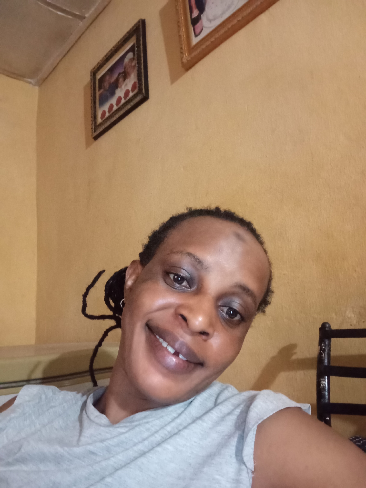 A person with dark skin and hair pulled back into twists, wearing a light gray T-shirt, is smiling in a selfie. The background shows an interior room with mustard-yellow walls. Two framed pictures hang on the wall. A bed is partially visible on the left, and a black chair is on the right. The location is likely Narayi, Nigeria, though this can't be confirmed with certainty.
