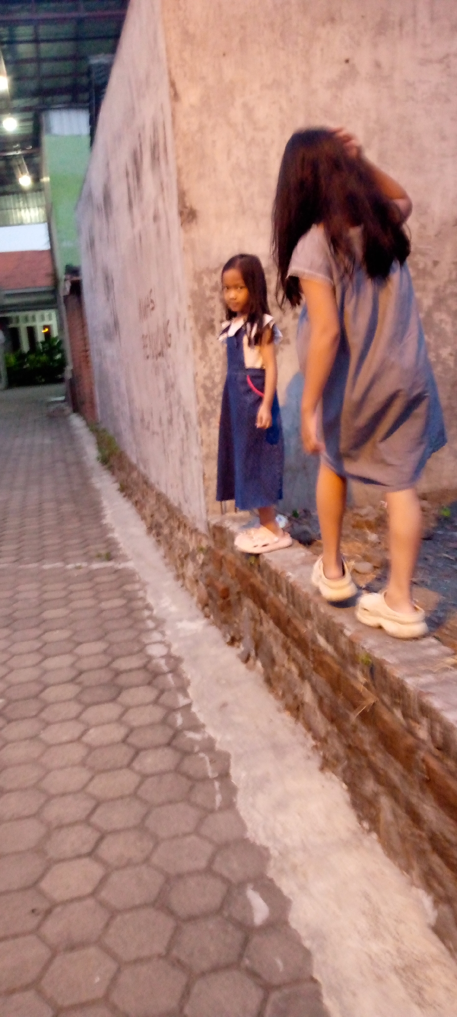 Two individuals, an adult and a child, stand on a low brick wall beside a concrete wall with graffiti in Sumberwono, Indonesia. The child, wearing a blue denim dress over a cream-colored top, faces the camera with her hands clasped in front of her. Her dark hair is slightly disheveled. The adult, seen from behind, has long dark hair and wears a gray t-shirt dress. They are both barefoot. The ground in front of them is a patterned concrete walkway, and behind them is a building with a corrugated roof. The lighting suggests it is dusk or evening.