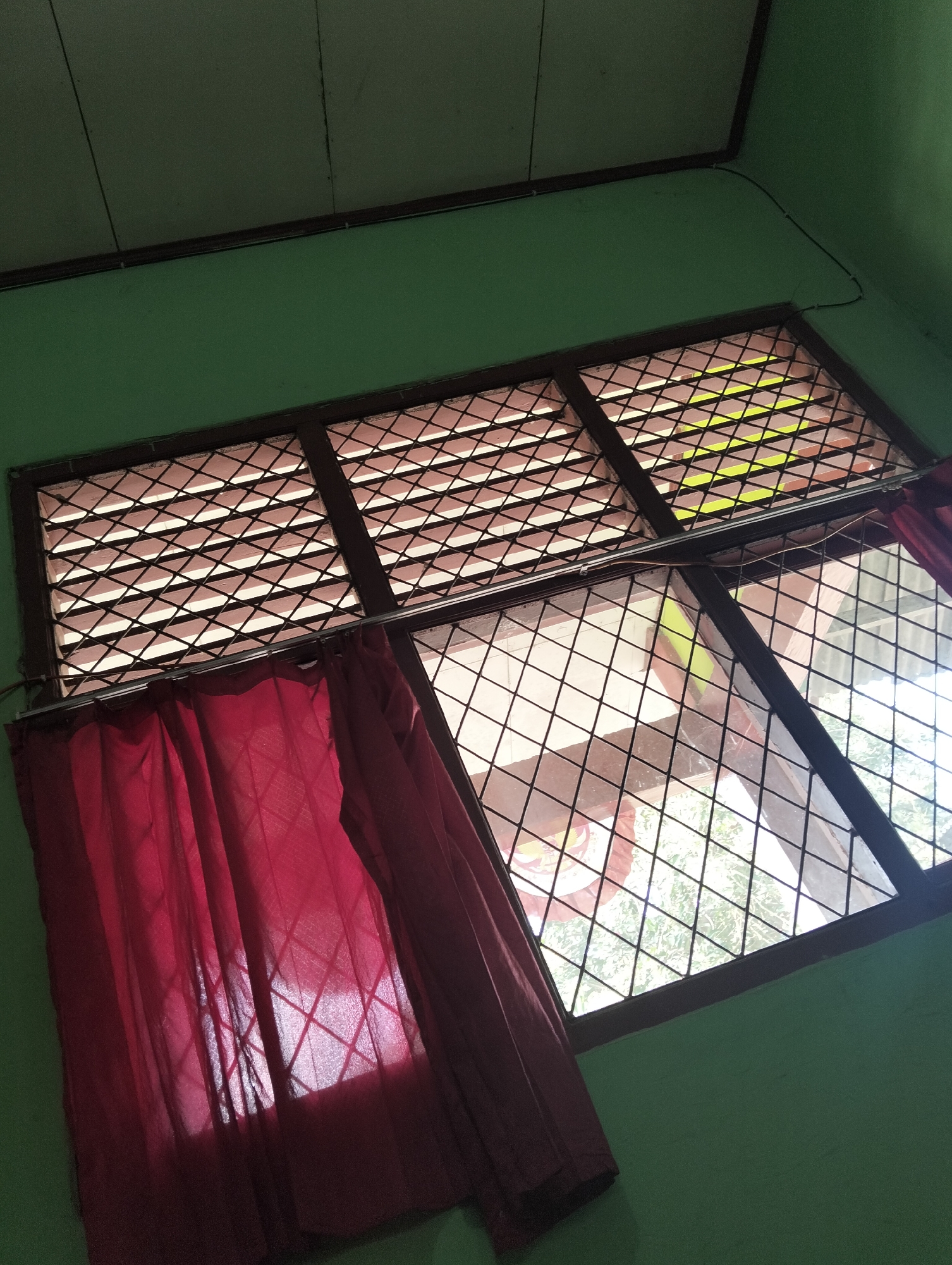 The image shows a window with a criss-cross metal grate and a red curtain partially covering it. The window is set into a green wall. The ceiling above is visible and appears to be made of wooden planks. Light filters through the window, casting shadows on the curtain. The location is identified as Banjar Agung, Indonesia. There are no people or specific actions depicted in the image.