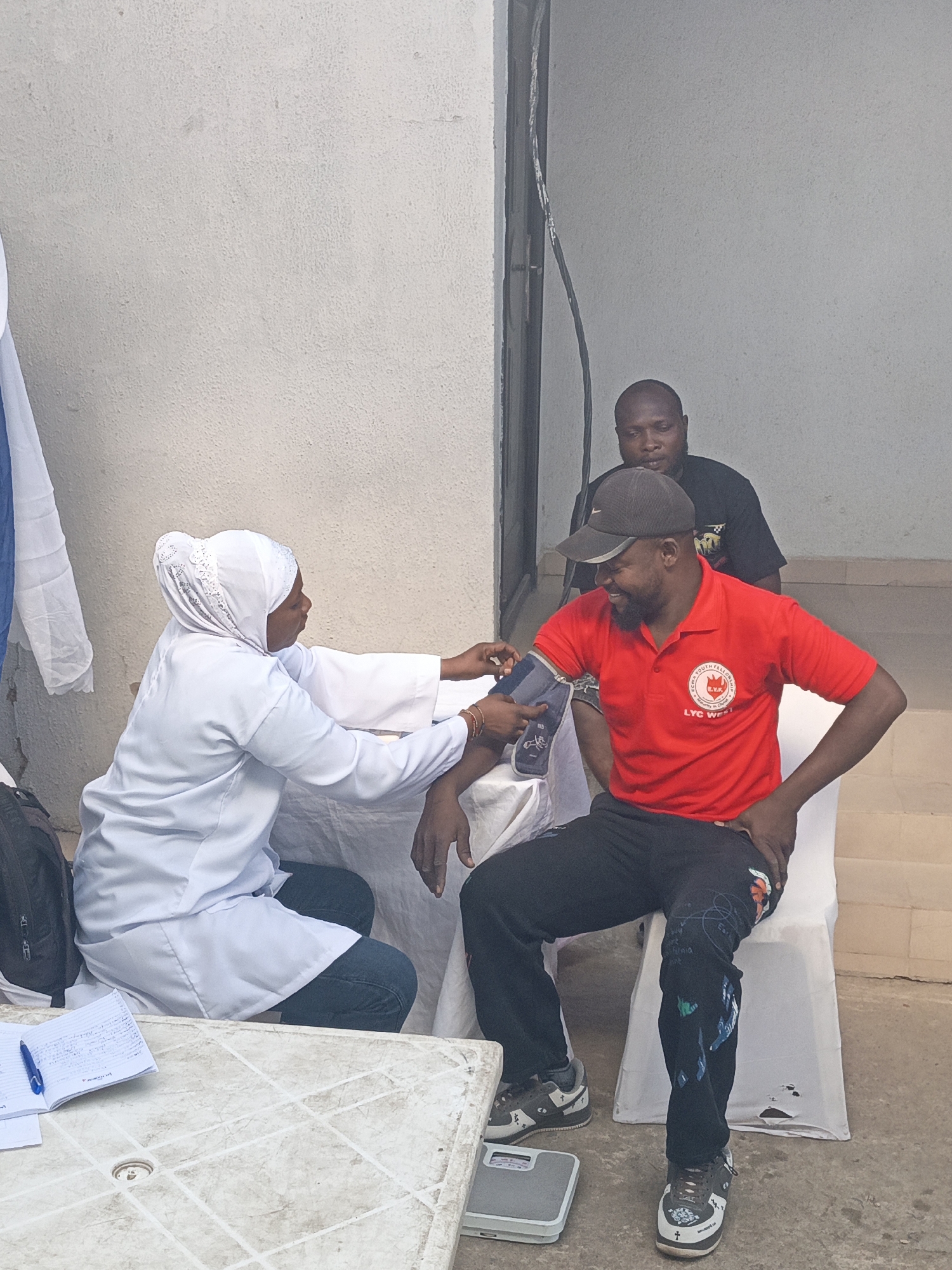 The image depicts a health screening or medical check-up taking place in a semi-outdoor setting, possibly against a building in Tarauni, Nigeria, during the daytime. The central activity shows a woman, dressed in a white lab coat and a white hijab, crouching to operate a blood pressure monitor on the left arm of a seated man.
The man, who appears to be the patient, is wearing a red polo shirt, a black baseball cap, and dark pants with abstract designs. He is seated on a white plastic chair and has a relaxed, smiling expression. His red polo shirt prominently displays a circular logo with the text "EKRA YOUTH FELLOWSHIP" and "LYC WEST" below it. On the ground next to his feet, a grey weighing scale is visible. His sneakers have white crosses and other designs.
Behind the man getting his blood pressure checked, another man in a black t-shirt is observing the process. To the left of the scene, a white plastic table holds an open notebook with handwritten notes and a blue pen. A black backpack is placed near the table. A blue and white piece of cloth hangs on the far left. The background is a plain, light-colored wall. The lighting suggests fair weather conditions during the day.