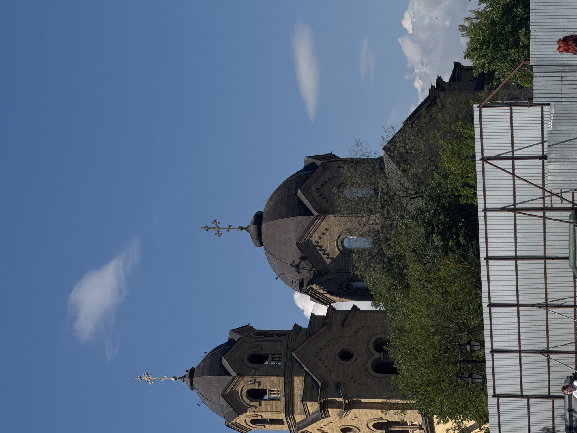 The image shows a vertically-oriented view of the Saint Nicholas Cathedral in Yevpatoriya, Ukraine. Two domed towers with crosses on top are visible against a blue sky with sparse white clouds. Green trees partially obscure the structures. To the right, there's a corrugated metal fence.