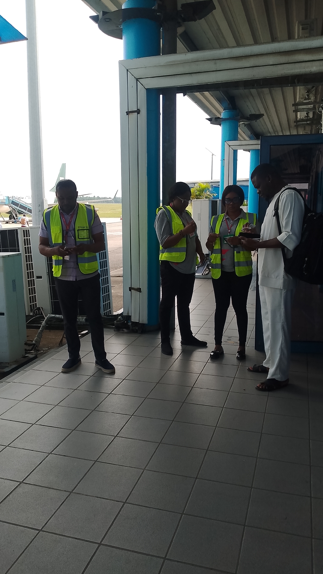 The image shows four people standing under a covered walkway with blue support columns and a tiled floor. Three of the people are wearing yellow-green safety vests, while the fourth person is wearing a white outfit. All of them appear to be looking at or holding some sort of card or phone. In the background, there is an airport runway, with a plane visible in the distance. Based on the limited visual data available, identifying the exact location within the city of Orerokpe, Nigeria, is not possible. The setting is consistent with an airport terminal or apron area, possibly for aircraft ground support or personnel.