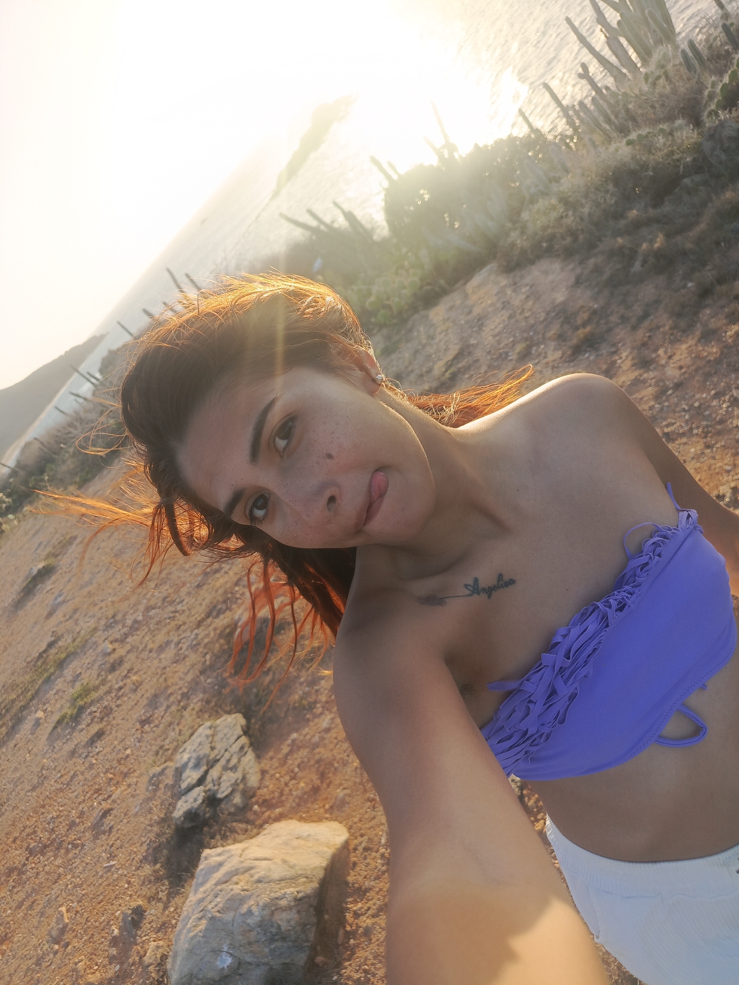 In Altagracia, Venezuela, a woman with reddish-brown hair takes a selfie at the beach. The sun is low in the sky, casting a warm glow and creating lens flare. She is wearing a purple bikini top with fringe detailing and white shorts. A tattoo reading "Angelica" is visible on her chest. She playfully sticks her tongue out at the camera. The background shows a sandy shoreline, scattered rocks, and cacti, with the ocean and distant hills visible in the distance. The image is taken from a low angle, looking up at the woman.
