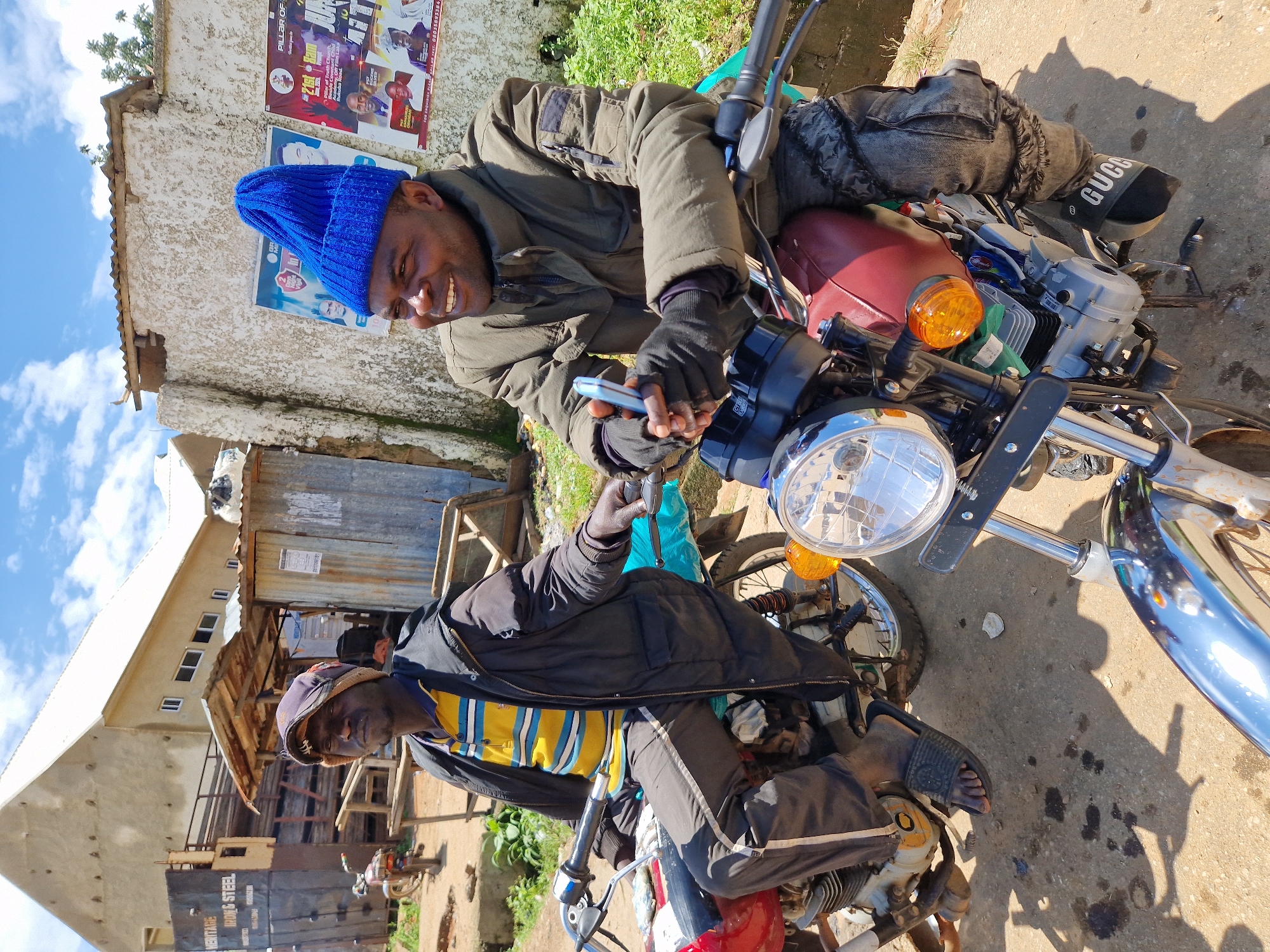 Two men are seated on motorcycles on a street in Kabong, Nigeria. The man on the left is wearing a yellow and blue striped shirt, a dark jacket, and a patterned beanie. The man on the right is wearing a green jacket, black gloves, and a blue knitted beanie. He is holding a silver mobile phone in his right hand and smiling. Both men are sitting on red and white motorcycles. The background shows a building, some corrugated metal structures, and posters on a wall. The ground is dirt and asphalt with some oil stains. The sky is blue with scattered clouds.