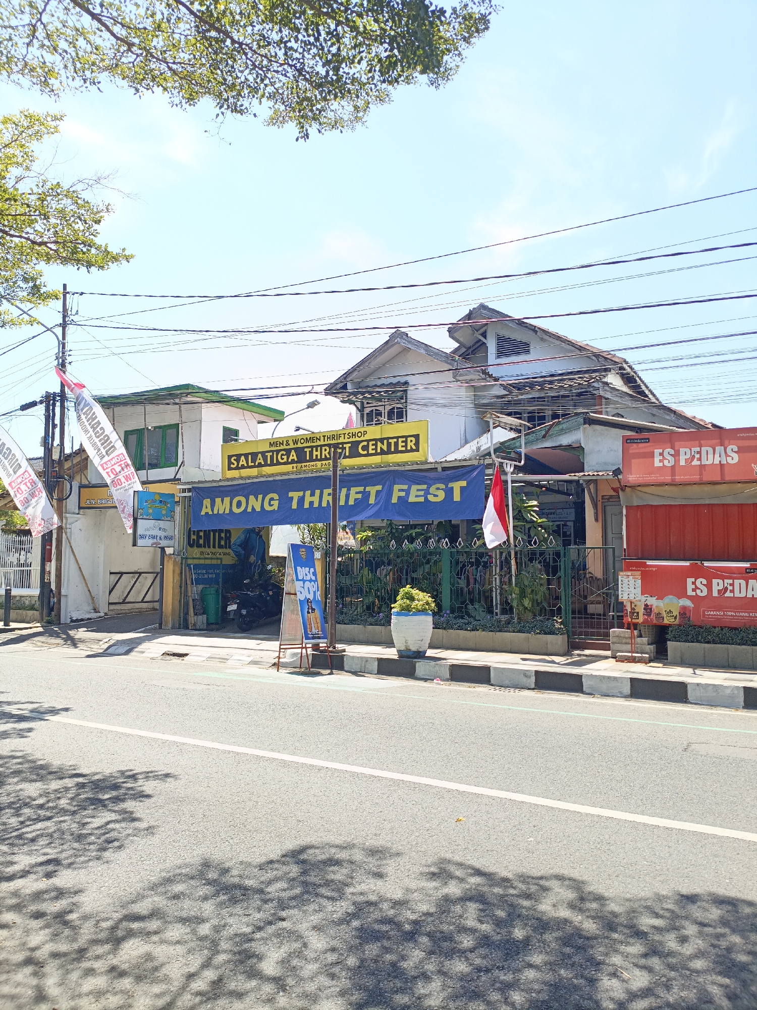 The image shows a street scene in Salatiga, Indonesia, on a sunny day. In the foreground, a paved road with a white line marking lanes is visible, with dappled shadows from trees falling across it. A concrete sidewalk with curb and planters runs alongside the road. Several buildings line the sidewalk.
The building on the left is a two-story structure with a white facade and green trim on the windows. A sign above the entrance reads "JLN. BRIGJEND SUDIARTO". Two white flags with red lettering are attached to a pole in front of this building.
Next to it is a single-story building with a green roof and white walls. A yellow sign on this building says "MEN & WOMEN THRIFT SHOP SALATIGA THRIFT CENTER BY AMONG PASAR". A blue banner stretches across the front of this building, with white text that appears to read "AMONG THRIFT FEST CENTER". An Indonesian flag, red and white, is displayed on a pole in front of the buildings.
To the right of the thrift shop, there are two red shipping container-like structures, one stacked on top of the other. Signs on these structures read "ES PEDAS". The lower container has graphics of drinks and text indicating "GARANSI 100% UANG KEMBALI".
A person wearing a blue jacket is standing near a black motorcycle in front of the thrift shop. A display stand with a poster showing "DISC 50%" is positioned on the sidewalk. A potted plant in a white pot with blue trim sits near the curb. Overhead, power lines crisscross the sky, and the tops of trees with green leaves are visible. The sky is bright blue with some wispy clouds.