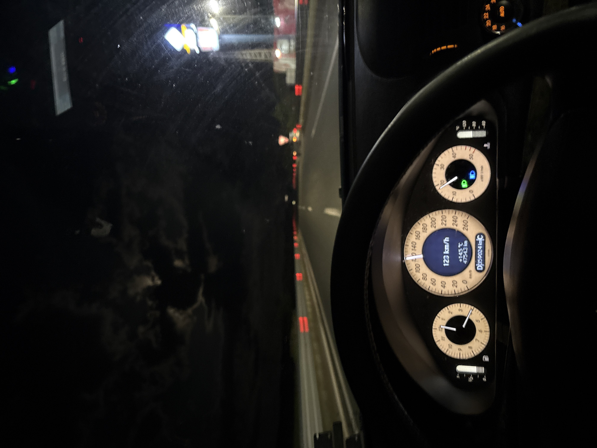 The image displays a night scene captured from the interior of a vehicle, viewed through the windshield towards the road ahead. The perspective suggests it was taken from the driver's or front passenger's seat.
**Subjects:** The foreground is dominated by the vehicle's dashboard and instrument cluster, partially framed by the steering wheel on the right. Outside, the road and distant vehicle lights are visible, along with glimpses of dark clouds and moonlight in the upper left portion of the external view.
**Activity or event taking place:** The vehicle is actively being driven at night.
**Notable details:** It is clearly nighttime, with the surroundings cloaked in darkness, illuminated only by artificial lights from the vehicle's interior and external sources. The instrument cluster shows the vehicle traveling at "123 km/h". The ambient temperature is displayed as "+14.5 °C", and a trip meter indicates "4754.3 km". The total odometer reads "D359024 kmC". A green indicator light confirms that the dipped beam headlights are active, and another green padlock symbol is illuminated. A display above the steering wheel shows "AUTO". Reflections are visible on the windshield, possibly from internal lights or dirt. The road ahead features red tail lights from other vehicles in the distance, implying traffic on what appears to be a highway or major road. Faint moonlight filtering through dark clouds adds to the nocturnal atmosphere. The context provided suggests this scene is in Dragichevo, Bulgaria.
**Visible text:**
- "123 km/h" (speed)
- "+14.5 °C" (temperature)
- "4754.3 km" (trip distance)
- "D359024 kmC" (odometer)
- "km/h" and "x1000 r/min" (gauge labels)
- Numerical scales on gauges (e.g., 0, 20, 40... 260 for speedometer; 0, 1... 6 for tachometer)
- "AUTO" (on climate control display)