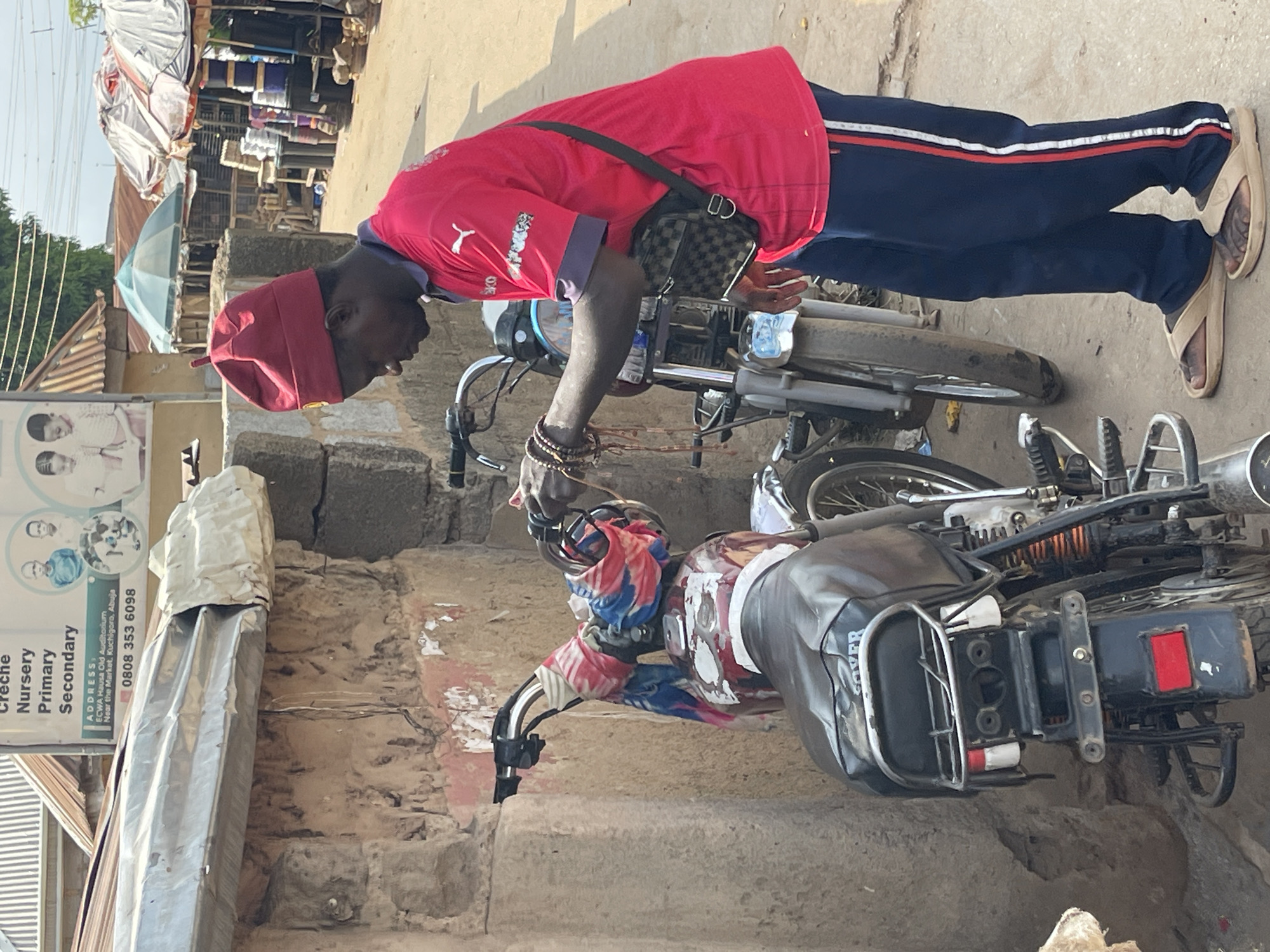 An outdoor scene in Kuchigoro, Nigeria, depicts a man leaning over a motorcycle on what appears to be a street or market area under bright daylight. The man is wearing a red long-sleeved shirt with a PUMA logo, dark blue track pants with red and white stripes, and beige flip-flops. He has a red cap, a checkered shoulder bag across his body, and multiple bracelets on his left wrist.
He is focused on a slightly worn motorcycle with a black seat, a red and white patched body, and handlebars adorned with colorful fabric. He appears to be inspecting or working on the motorcycle's front section, holding a small clear bottle in his right hand. A small water bottle rests on the motorcycle's fuel tank. A second motorcycle is partially visible behind him.
The background features various structures, including buildings or stalls, some covered with tarpaulin, and a light blue and white striped umbrella. The ground beneath is paved or dirt.
A prominent sign on the left side of the image provides educational and contact information, reading "Nursery Primary Secondary" and "ADDRESS: ECWA Hausa Old Auditorium Near the Market, Kuchigoro, Abuja," along with a phone number "0808 353 6098." The words "Эко" and "CO" are also visible on the sign, likely part of a logo. The word "SILVER" is visible on the motorcycle's seat or side panel.
