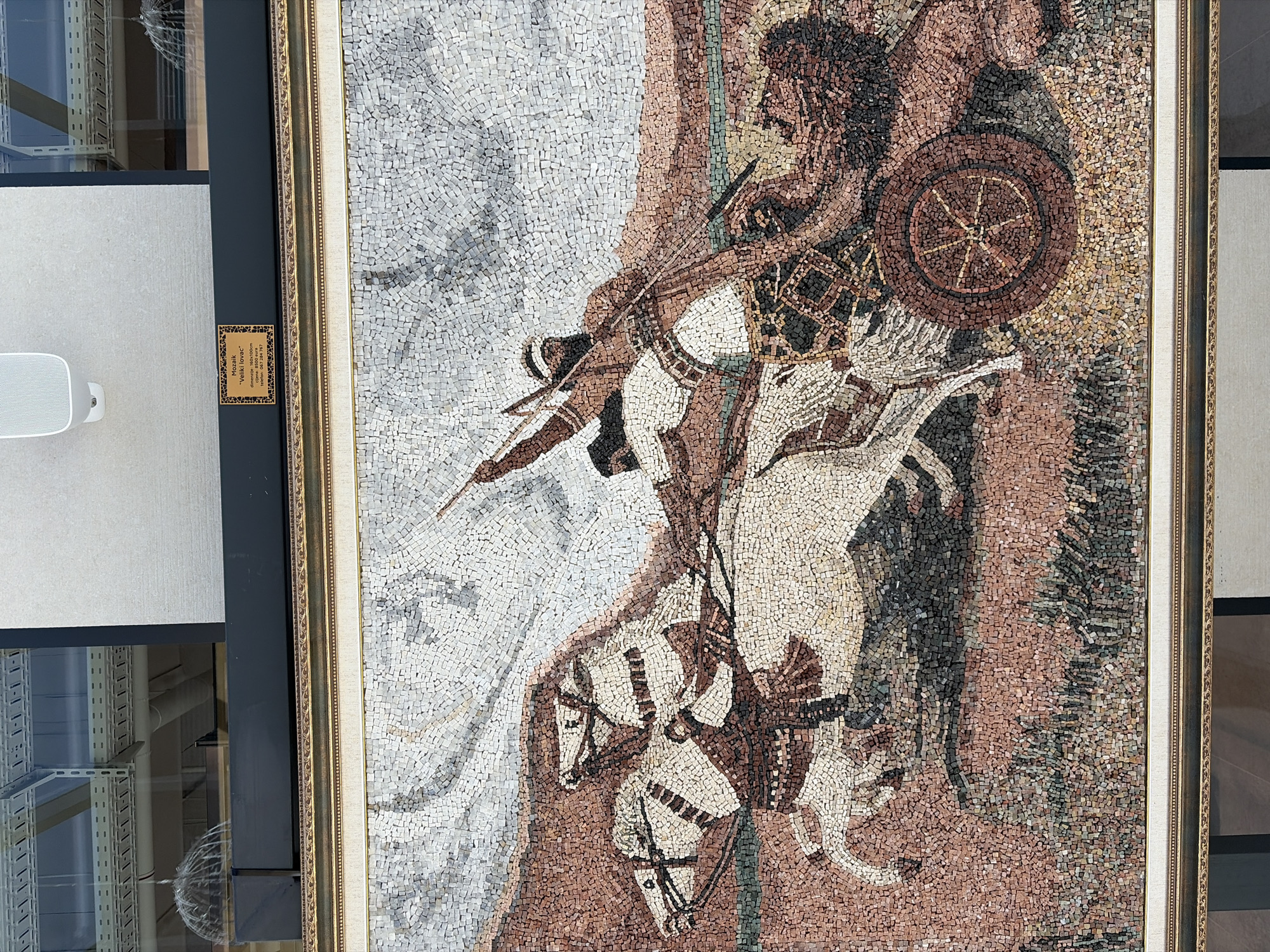 The image shows a large mosaic depicting a scene with a chariot pulled by two horses. A figure in the chariot holds a spear and appears to be in motion, possibly hunting or in battle. The mosaic is framed in gold and is displayed in a museum or gallery setting. A small plaque to the upper right of the mosaic reads "Mezik \"Veliki lovac\" dimenzije 160x100cm cijena 8500 km tel. +387 65 504 787". This translates to "Mosaic \"The Great Hunter\" dimensions 160x100cm price 8500 km tel. +387 65 504 787". This artwork is on display at the National Museum of Montenegro in Budva, Montenegro.