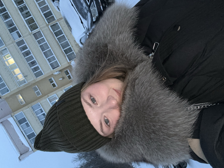 A person wearing a dark green beanie and a black winter coat with a large grey fur collar looks towards the camera. The background features a light yellow building with rows of windows and snowy ground. The photograph is taken from an unusual angle, slightly tilted. The scene suggests a cold, possibly snowy environment.