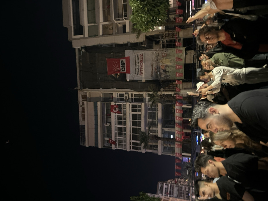The image shows a gathering of people in the evening in İzmir, Türkiye, at the coordinates provided. The crowd is standing in front of a building that is partially obscured by a large banner. The banner is red and white with the letters CHP in large, bold letters, followed by the text "Aday yarımada sandığı bizimde İSTİYORUZ." The banner is hanging from the building's second floor, and there are several Turkish flags draped across the building. The crowd is facing the building, and many are holding their cell phones up, possibly taking photographs or videos. There are palm trees visible in the background, and the street is lined with buildings, some of which are lit up. The scene appears to be a political rally or demonstration. The crowd is diverse in age and gender, with a mix of men and women. The people in the foreground are close to the camera, with some faces obscured by others. The scene is lit by streetlights and the ambient glow of the city lights, which creates a soft and hazy effect.
