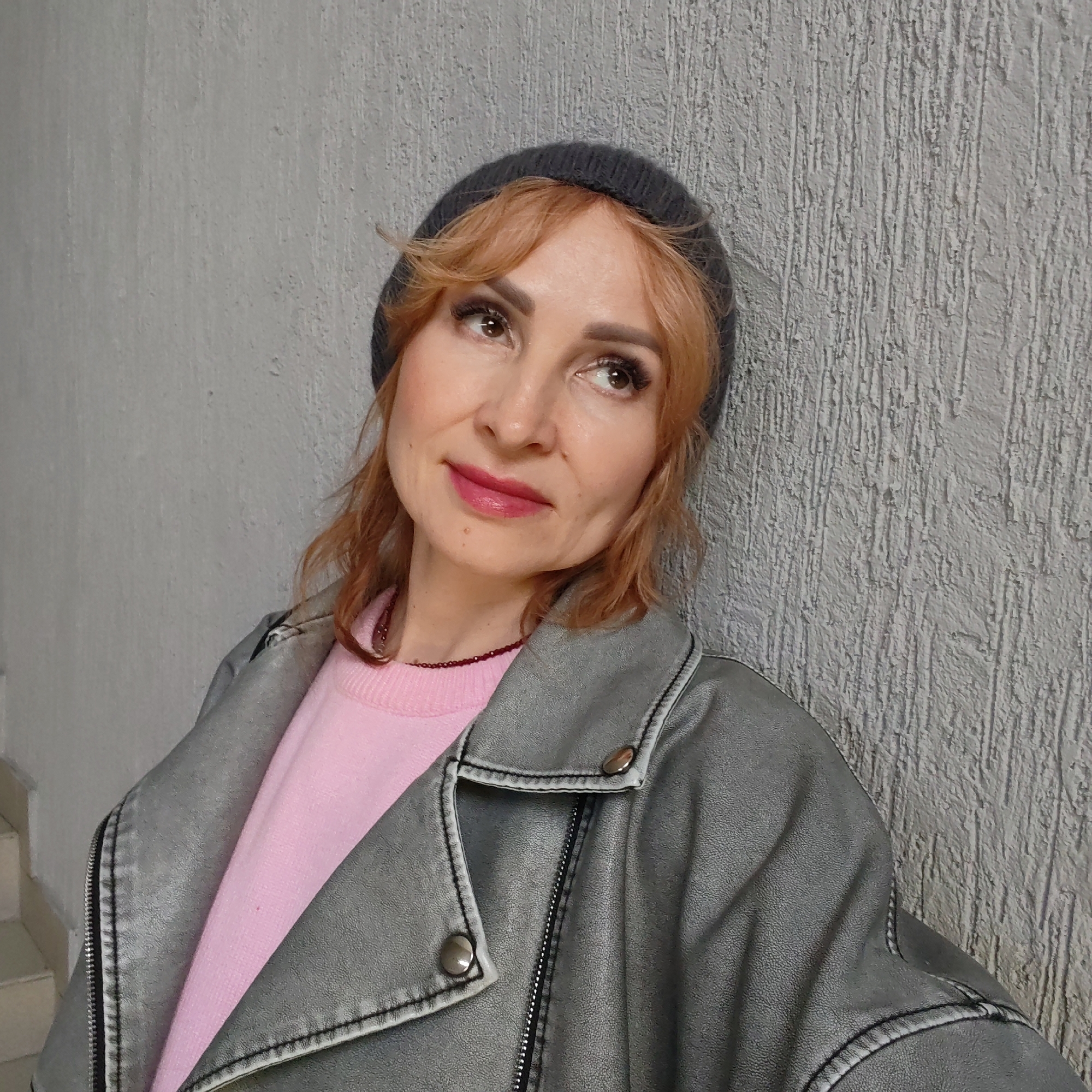A woman leans against a textured, light gray wall. She is wearing a dark gray beanie, a pink sweater, and a distressed gray leather jacket. She is looking upwards with a slight smile. A beaded necklace is visible at her neck. Part of a staircase is visible in the lower-left corner of the image. The location is Chaikovsky, Russia.