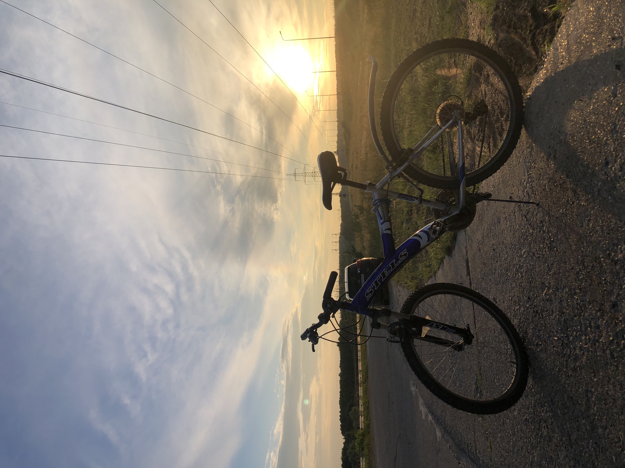 The image shows a bicycle leaning on the side of a paved road, with its wheels resting on the asphalt and grass. The bicycle is blue and white and has the brand name "STELS" visible on the frame. The scene is set outdoors during sunset, as indicated by the warm, golden light and the long shadows cast by the bicycle and surrounding objects. The sky is partly cloudy, with streaks of white clouds against a backdrop of pale orange and yellow hues from the setting sun.
In the background, power lines stretch across the sky, supported by tall poles and a large lattice tower. Beyond the power lines, a treeline is visible on the horizon, suggesting a rural or semi-rural setting. Several streetlights are also present along the road, illuminated by the fading daylight. The overall mood of the image is serene and peaceful, with a sense of quiet solitude.
The orientation of the image is vertical, with the sun appearing at the top left of the frame. The time of day is clearly late afternoon or early evening, as the sun is low in the sky and casting a warm, diffused light. The weather appears to be clear, with no strong winds or rain, given the stillness of the clouds and the grass. The location, Chaikovsky, Russia, is a city in Perm Krai, known for its industrial and natural landscapes.