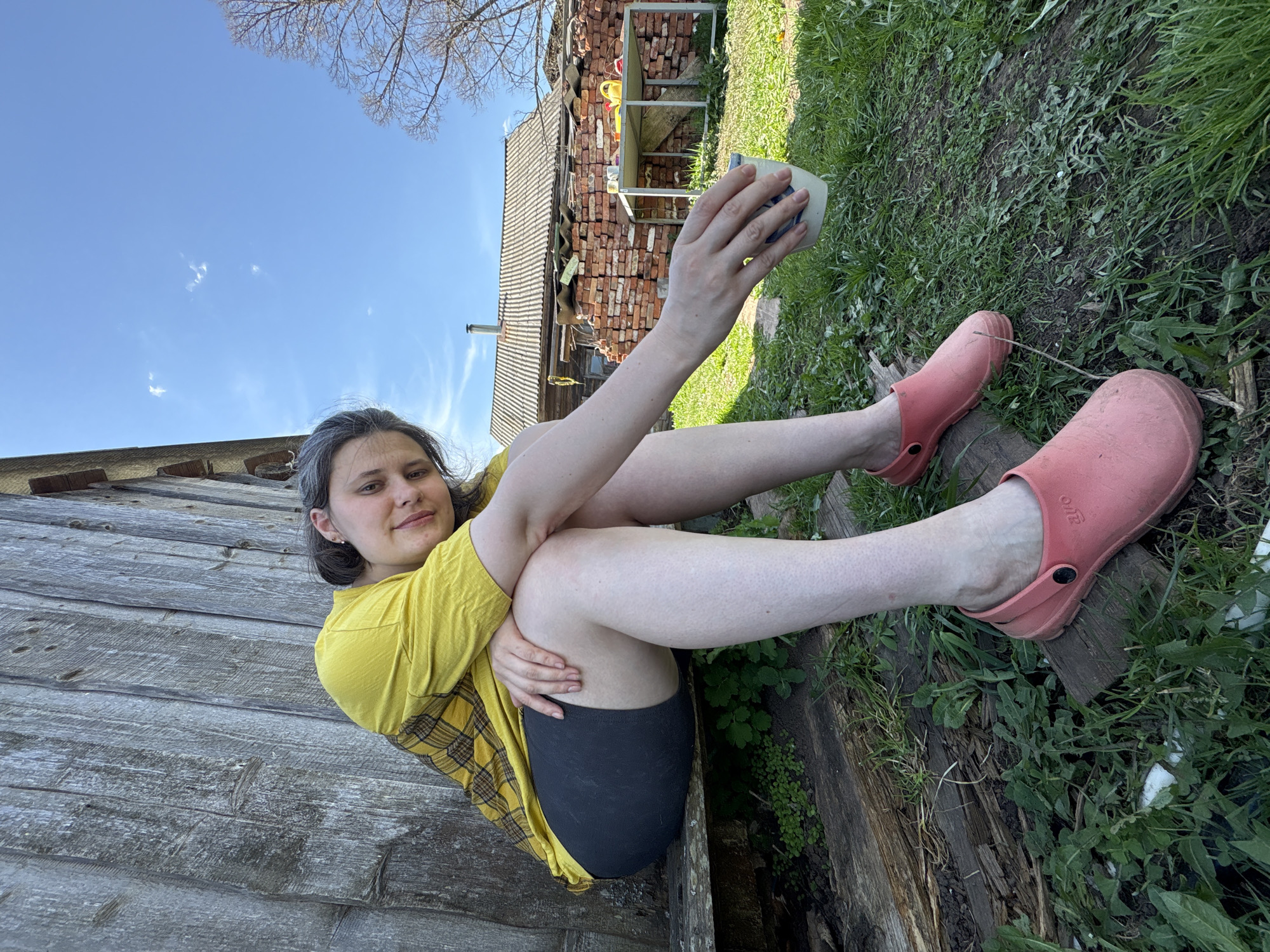 A young woman is sitting outdoors in Nizhnyaya Chermoda, Russia. She is wearing a yellow t-shirt with a yellow and black plaid shirt over it, black shorts, and pink clogs. She is holding a white and blue mug in her hand. She is sitting on a wooden structure, possibly a fence or a bench. In the background, there is a house or shed with a brick pile next to it, grass, and a blue sky with some clouds.
