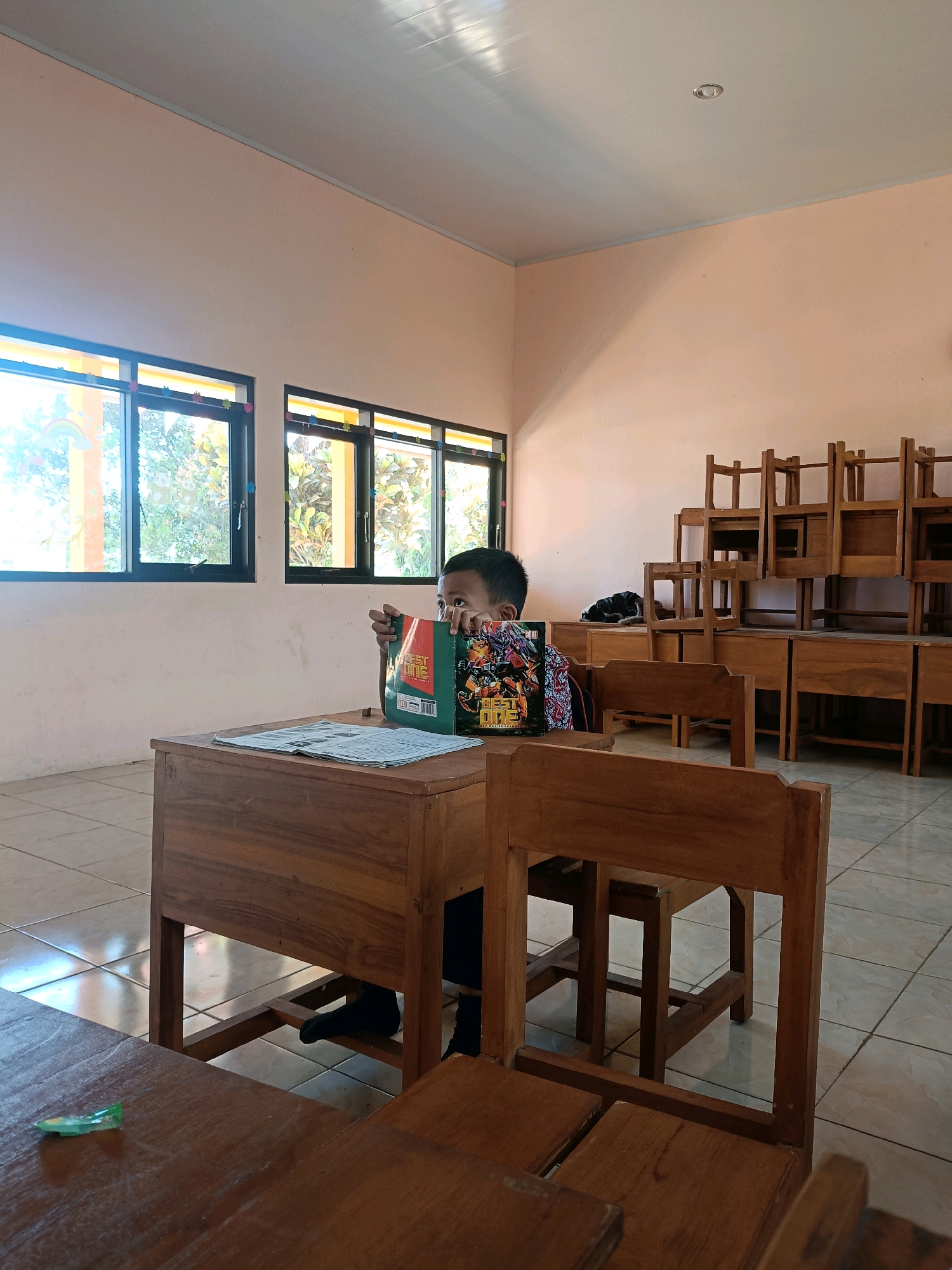 In a classroom located in Gunungwungkal, Indonesia, a young student is seated at a wooden desk. The student, partially obscured by a book, holds it in front of their face, suggesting an attempt to hide or avoid being seen. On the desk, in addition to the book, lies a newspaper. The classroom environment is furnished with rows of stacked wooden chairs and more wooden desks. Sunlight streams in through the windows, illuminating the tiled floor and the pale pink walls.