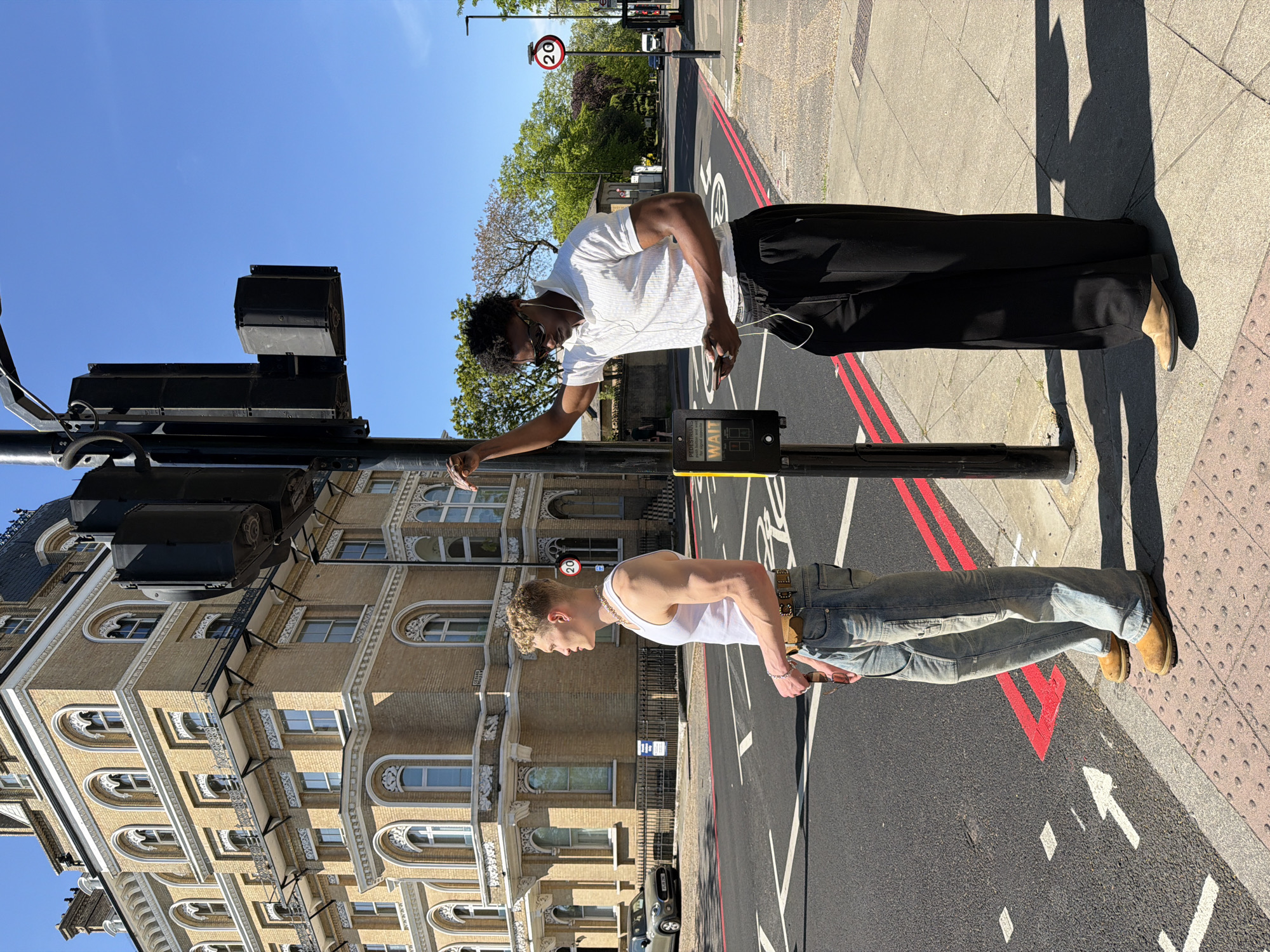 Two men stand at a pedestrian crossing in London, United Kingdom. One man, Black, wears a white t-shirt, black trousers, and tan shoes. He is looking at his phone and has headphones on. The other man, Caucasian, wears a white tank top, jeans, and tan shoes. He is also looking at his phone. They are at a pedestrian crossing on a street with red lines and white arrows painted on the asphalt. A traffic light pole is next to them, and a building with arched windows is in the background. A "20" speed limit sign is also visible.