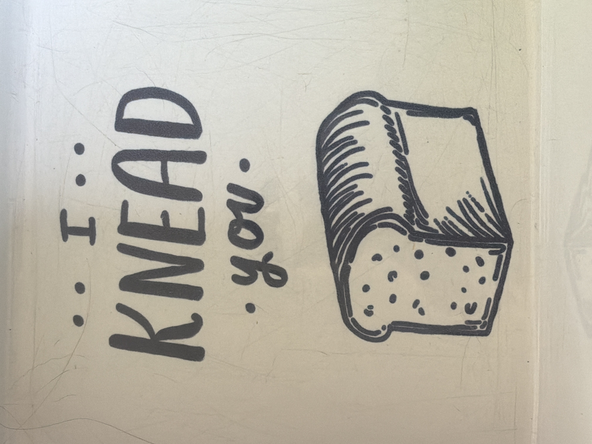 The image is a close-up, eye-level shot of a sign with a pun. The text reads "...I KNEAD you." written in a handwritten font. Below the text is a hand-drawn image of a slice of bread. The sign is off-white and has a slightly textured surface with some visible scratches and imperfections. The lighting is bright and even, casting no significant shadows. The background is a uniform light color, suggesting the sign is indoors or in a controlled environment. There is no information in the image to indicate its location in Portland, United States of America, or any details about people or specific actions.