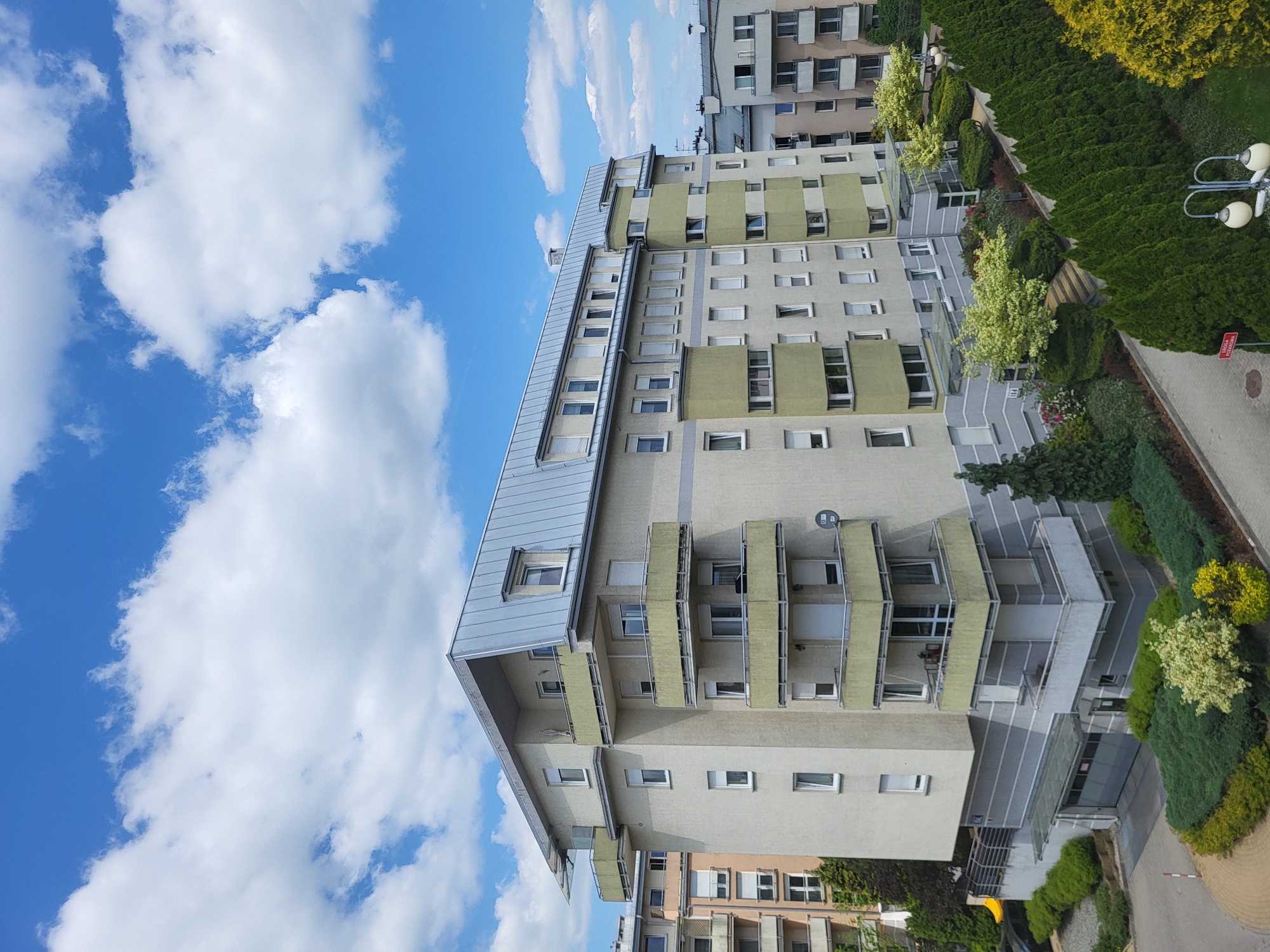 The image shows a multi-story apartment building in Unknown, Unknown. The building has a light beige facade with green accents and gray metal roofs on the upper levels. Balconies with metal railings are visible on several floors. To the right of the building, there is lush greenery, including shrubs and trees. A paved walkway or street is adjacent to the greenery. The sky is a bright blue with scattered white clouds. The building appears to be in a residential area with other similar buildings nearby.
