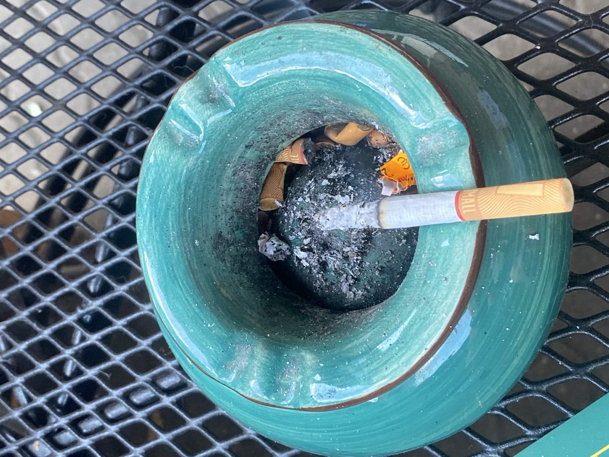The image shows an outdoor scene, likely on a patio table, with a green ceramic ashtray filled with cigarette butts and ash. A single cigarette rests in the ashtray, with its filter visible and the burning end partially obscured by ash. The ashtray is situated on a black metal mesh surface. The background is out of focus, but appears to be a continuation of the mesh surface, suggesting an outdoor furniture setting. The lighting suggests it is daytime, with natural light illuminating the scene. There is no indication of specific location within Lütjenwestedt, Germany, or any people present.