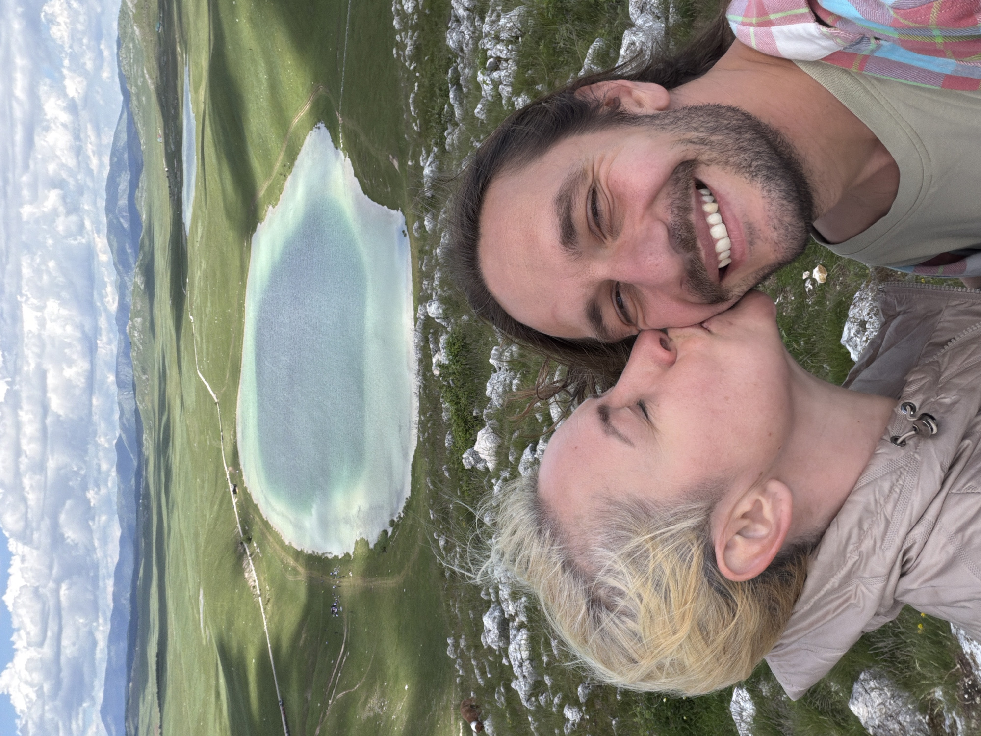 A close-up, selfie-style photograph captures two individuals embracing against a panoramic natural backdrop identified as Novakovići, Montenegro. In the foreground on the left, a woman with short blonde hair and fair skin has her eyes closed as she kisses the man's right cheek. She is dressed in a light-colored, possibly beige or khaki, quilted jacket with a high collar. On the right, a man with medium-length dark brown hair and a beard smiles broadly at the camera, his eyes open. He wears a light green t-shirt beneath an open plaid shirt featuring shades of red, blue, and white. The immediate ground around the individuals is rocky, covered with numerous small, light-grey stones interspersed with green grass.
The background presents an expansive, undulating landscape dominated by two lakes. The most prominent is a large, oval-shaped body of water in the mid-ground with strikingly clear, light turquoise-blue water. Its shores are lined by green grassy banks, with unpaved dirt roads or paths winding along its perimeter. A smaller, irregularly shaped lake is visible further back and to the right of the main lake. The lakes are nestled within vast, gently rolling green hills, indicative of pastures or meadows, which extend towards distant, hazy blue mountain ranges on the horizon. The sky above is partly cloudy, featuring significant white cumulus clouds against a lighter blue backdrop, with cloud shadows evident across the hills. A small gathering of individuals or objects appears near the shoreline of the main lake, adjacent to a dirt path.