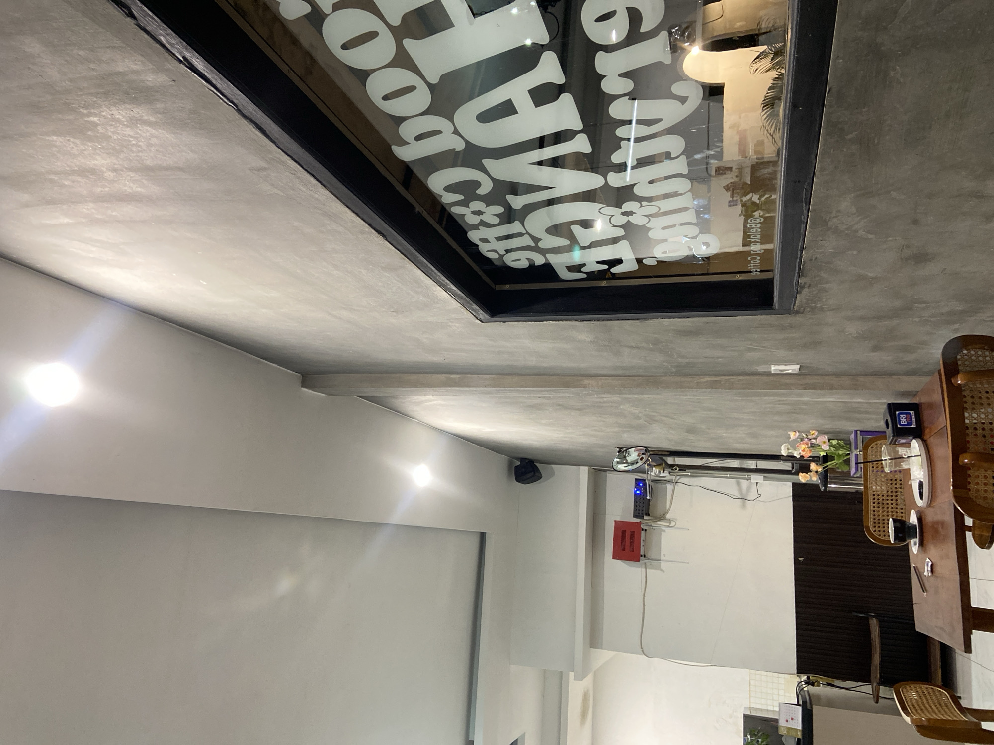 This image depicts the interior of a cafe, likely in Sekar Mawar, Indonesia. The scene is dominated by concrete and white walls, creating a minimalist and modern aesthetic. A large window with white text on it is a prominent feature, reflecting light and showing a glimpse of the outside. The text appears to be a logo or name for the establishment, with stylized lettering and floral elements.
In the foreground, a wooden table with chairs is set up, suggesting a dining or seating area. On the table, there is a coffee cup and saucer, a small vase with flowers, and what looks like a device with a blue light, possibly an electronic gadget. The lighting is a mix of bright artificial lights and natural light filtering in, creating reflections on the surfaces. The overall ambiance suggests a quiet, perhaps evening or late afternoon setting, as indicated by the indoor lighting. There are also some visible electronic devices mounted on the wall, including a red box and what appears to be a security camera. The image is taken from a slightly angled perspective, adding depth to the scene.