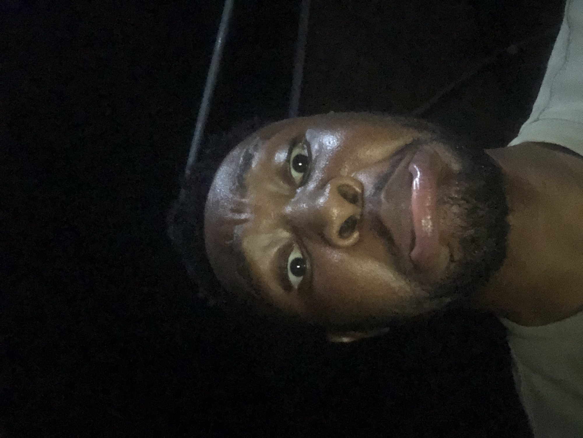 A close-up selfie of a Black man with dark hair and a beard. His skin appears oily or sweaty. The background is dark, with some blurred vertical bars visible at the top. He is wearing a light-colored shirt. The location is Unknown, Unknown.