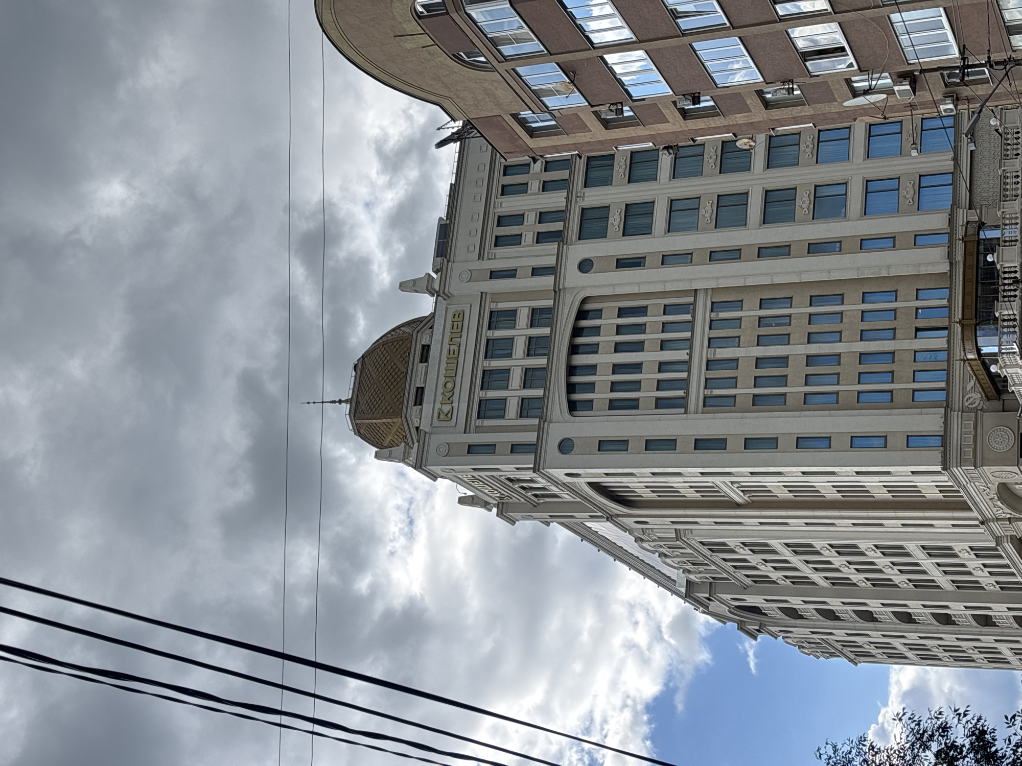 The image shows the "Koshelev" residential complex in Novokuybyshevsk, Russia, on an overcast day. The complex is a tall, multi-story building with a beige facade and a distinctive golden dome. The building features numerous windows, some with blue tinted glass, and decorative architectural elements. To the right, another building with a brown facade and balconies is partially visible. Overhead, black electrical wires crisscross the sky, contrasting with the grey clouds and patches of blue sky. The perspective is from below, looking up at the buildings. No people are visible in the image.