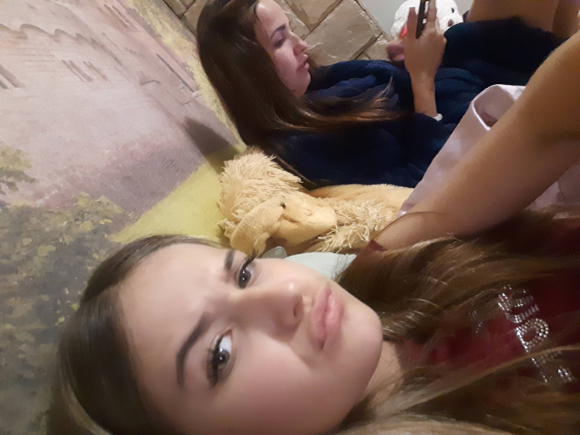The image features two young females lounging on a couch. The girl in the foreground is looking directly at the camera with a playful expression, her face filling a significant portion of the frame. She has long, brown hair, styled with some volume. The other girl, in the background, is lying down and looking at her phone, also with dark hair. She is wearing a dark blue robe. A fuzzy, light brown stuffed animal is visible between the two girls, and the couch appears to have a textured, light brown and yellow patterned backdrop. The lighting suggests it might be indoors during the evening or a dimly lit room.