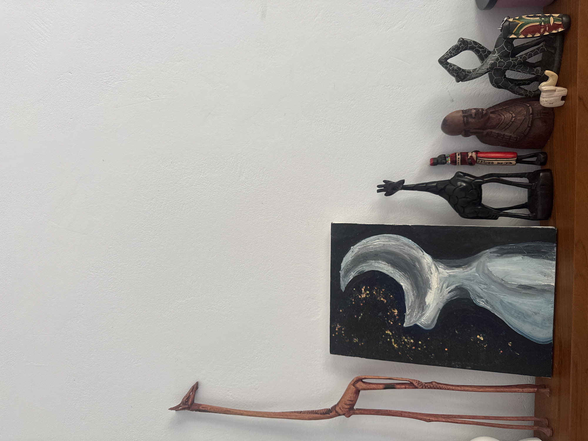 The image shows a white wall with a collection of wooden sculptures and a painting arranged vertically along its right side. The sculptures depict various figures, including stylized animals and human-like forms, possibly of African origin. The painting on canvas features a white, abstract shape on a black background with gold specks. The bottom wooden sculpture is a long-necked bird. The sculptures and the painting are attached to a wooden frame that runs vertically along the right side of the wall. The image was likely taken in Pejeng, Indonesia.